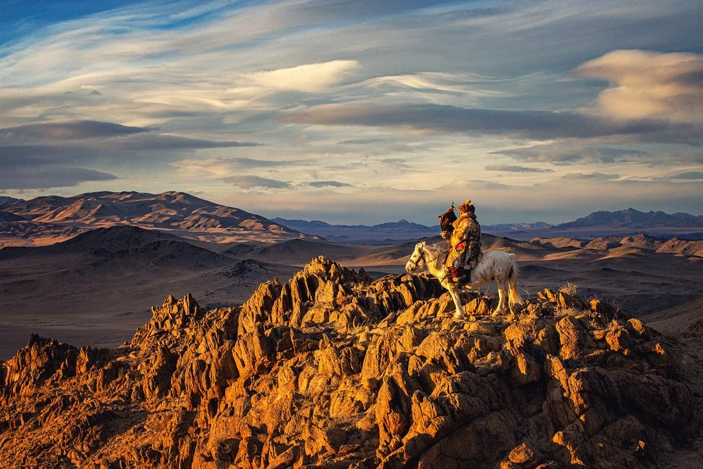 A person in traditional attire riding a white horse on a rocky hilltop in a vast desert landscape during sunset, with mountains and a partly cloudy sky in the background.