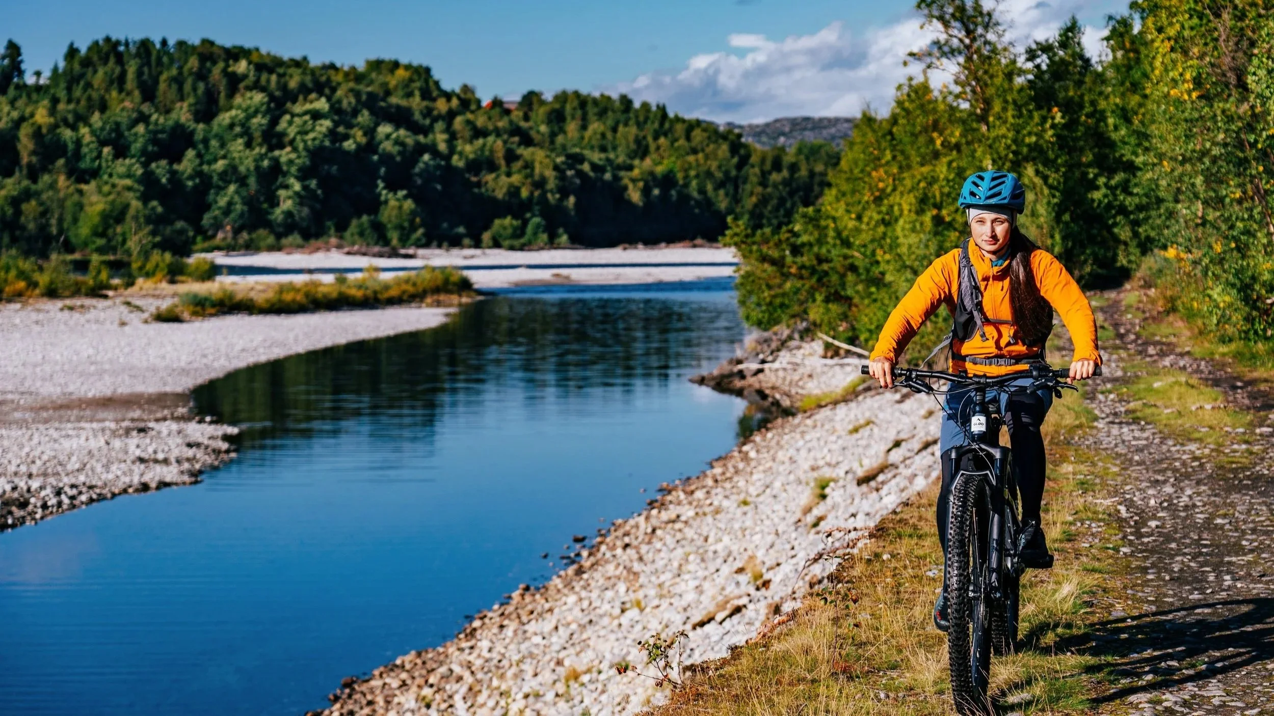 Woman in an orange jacket and blue helmet riding a mountain bike along a river trail surrounded by trees and hills.