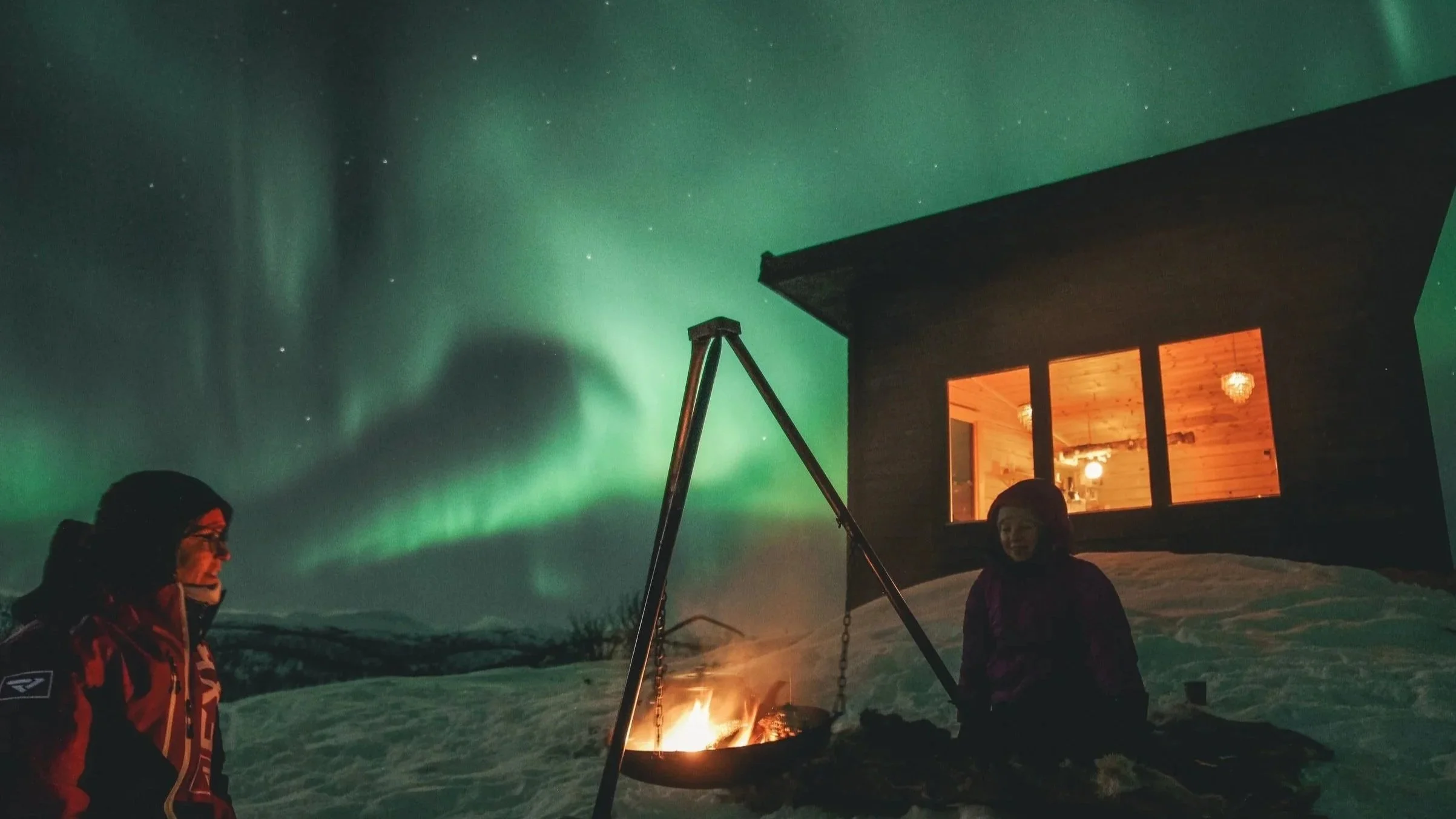 Two people sitting around a campfire outdoors at night, with a house behind them and the Northern Lights illuminating the sky.