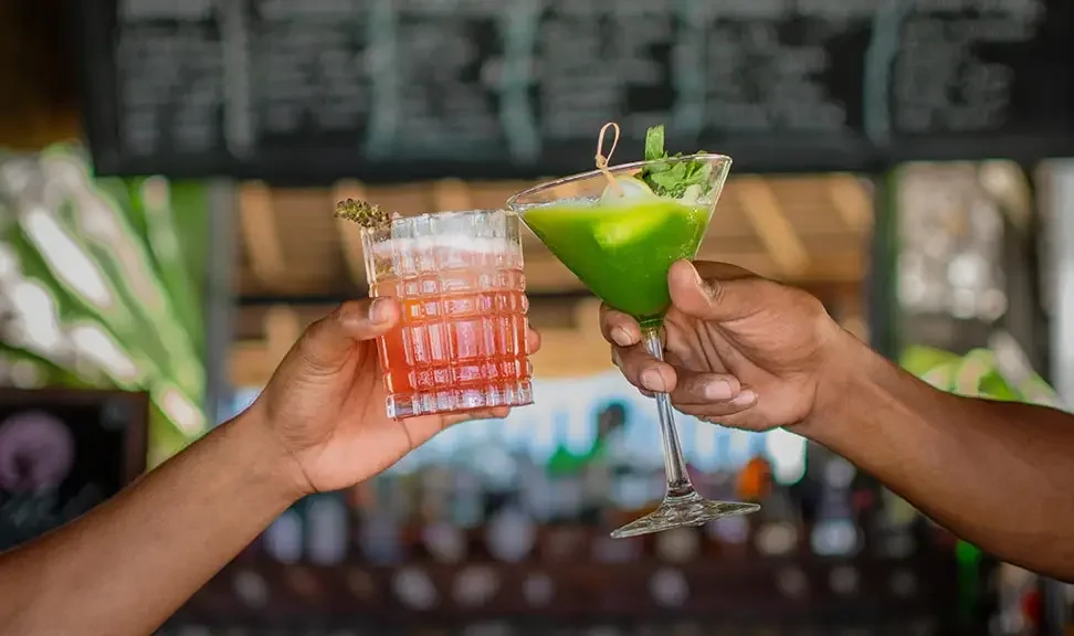 Two people clinking drinks, one in a textured glass with a pink-colored cocktail garnished with a flower, and the other in a martini glass with a green cocktail garnished with a lime wedge and mint.