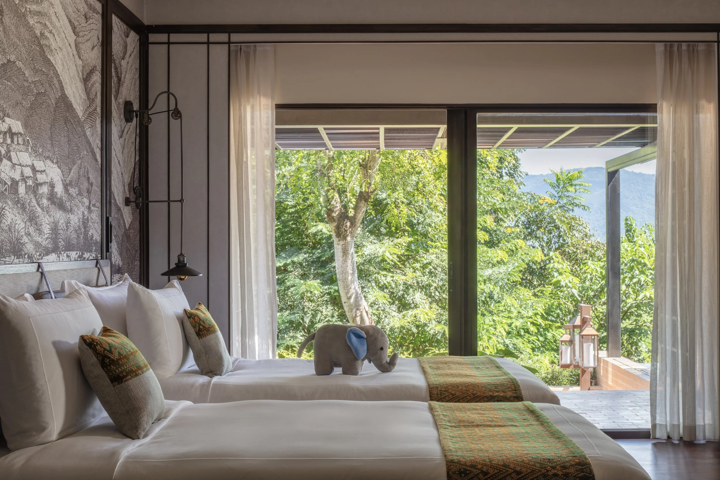 A bedroom with two beds, decorative pillows, and a stuffed elephant on one bed. Large glass doors lead to a balcony with lush green trees and hills in the background.