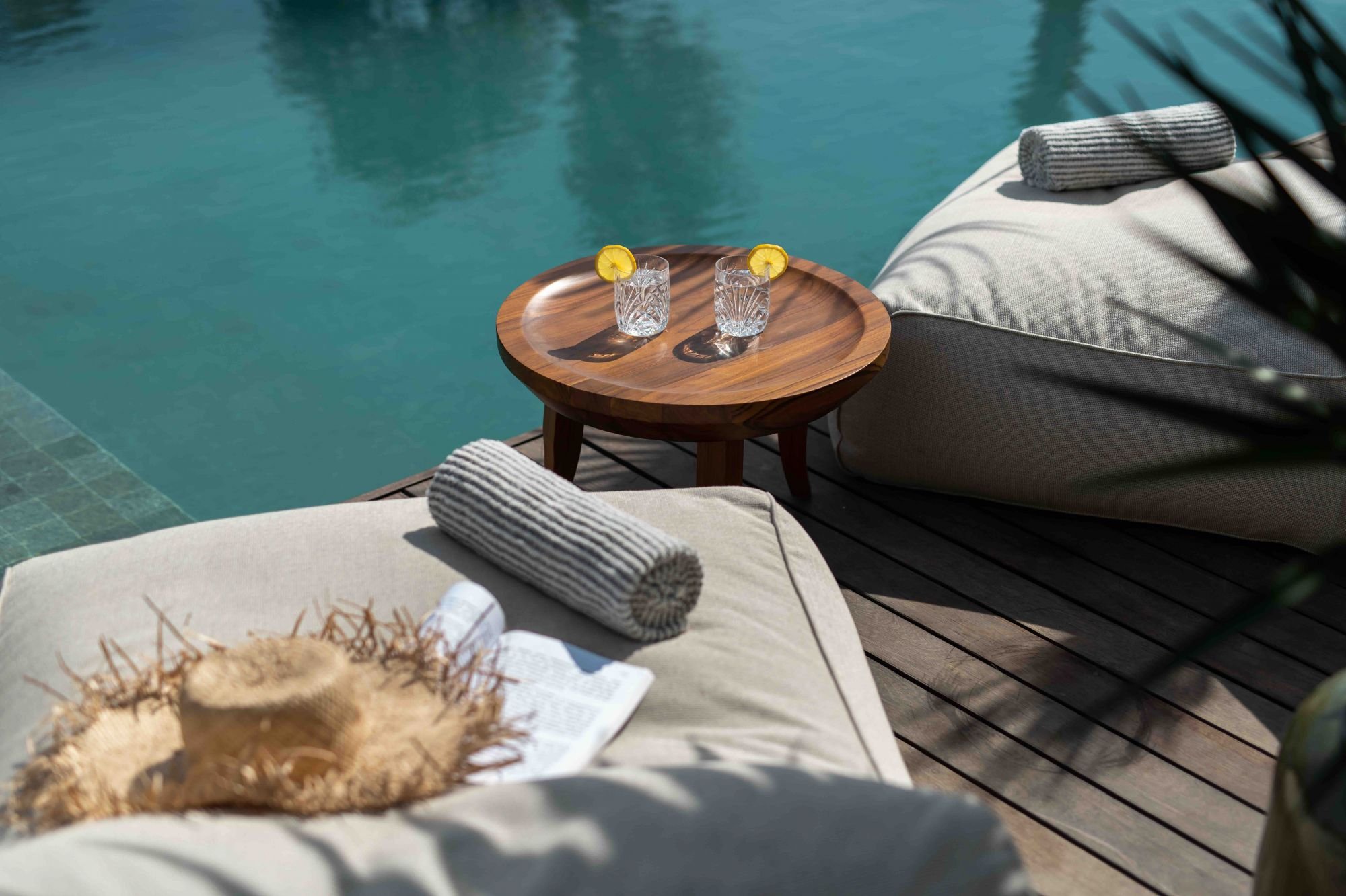 Luxury poolside lounge chairs with rolled towels, a straw hat, and a magazine on a Cushioned daybed beside a pool, with a small wooden table holding two glasses of water with lemon slices.