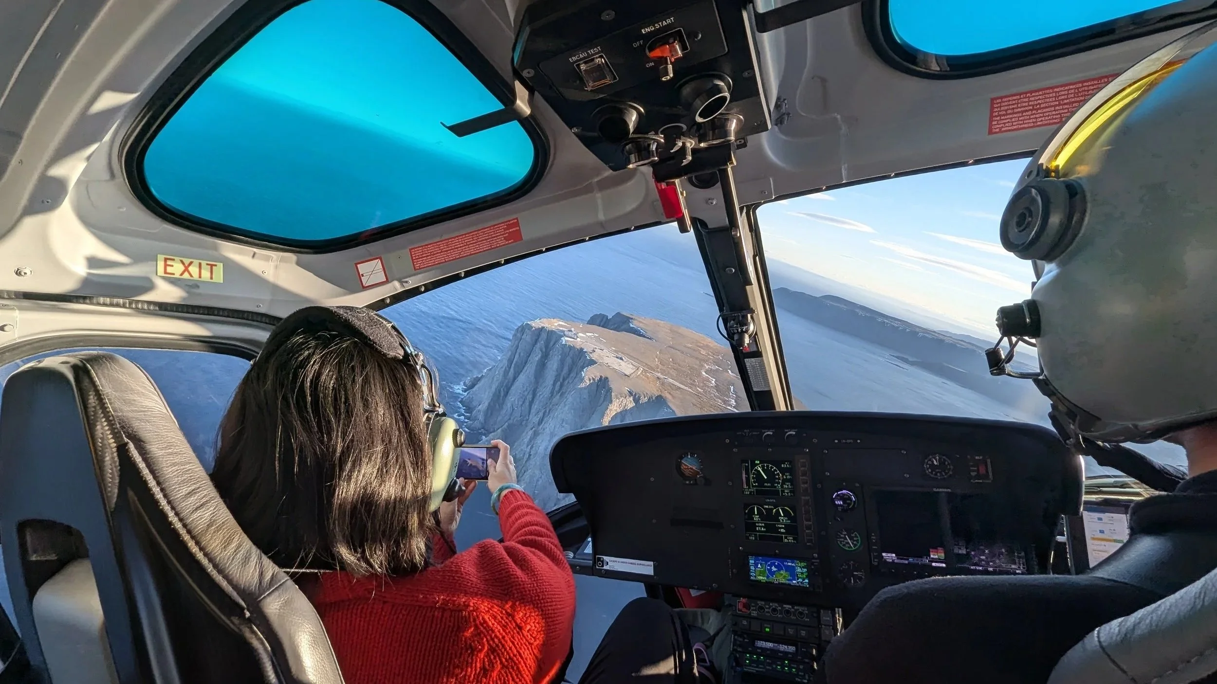 Inside an aircraft cockpit, a woman wearing a red sweater is taking a photo of the view outside the window with her phone. The pilot wearing a helmet is seated next to her. The cockpit shows various instruments and a large window revealing a coastline with cliffs, water, and a partly cloudy sky.