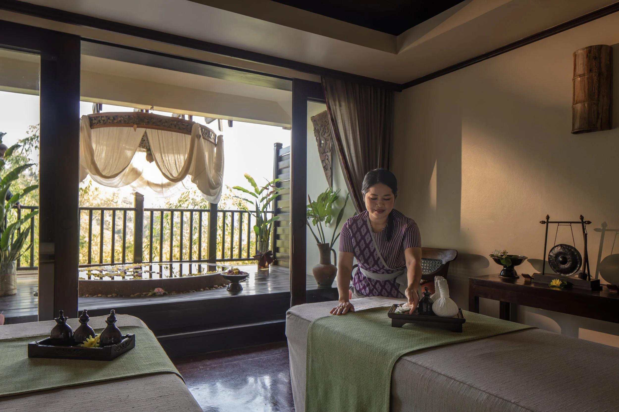 A woman in traditional attire preparing a massage table at a spa, with an open balcony door showing a decorated outdoor area with plants and a hanging canopy.