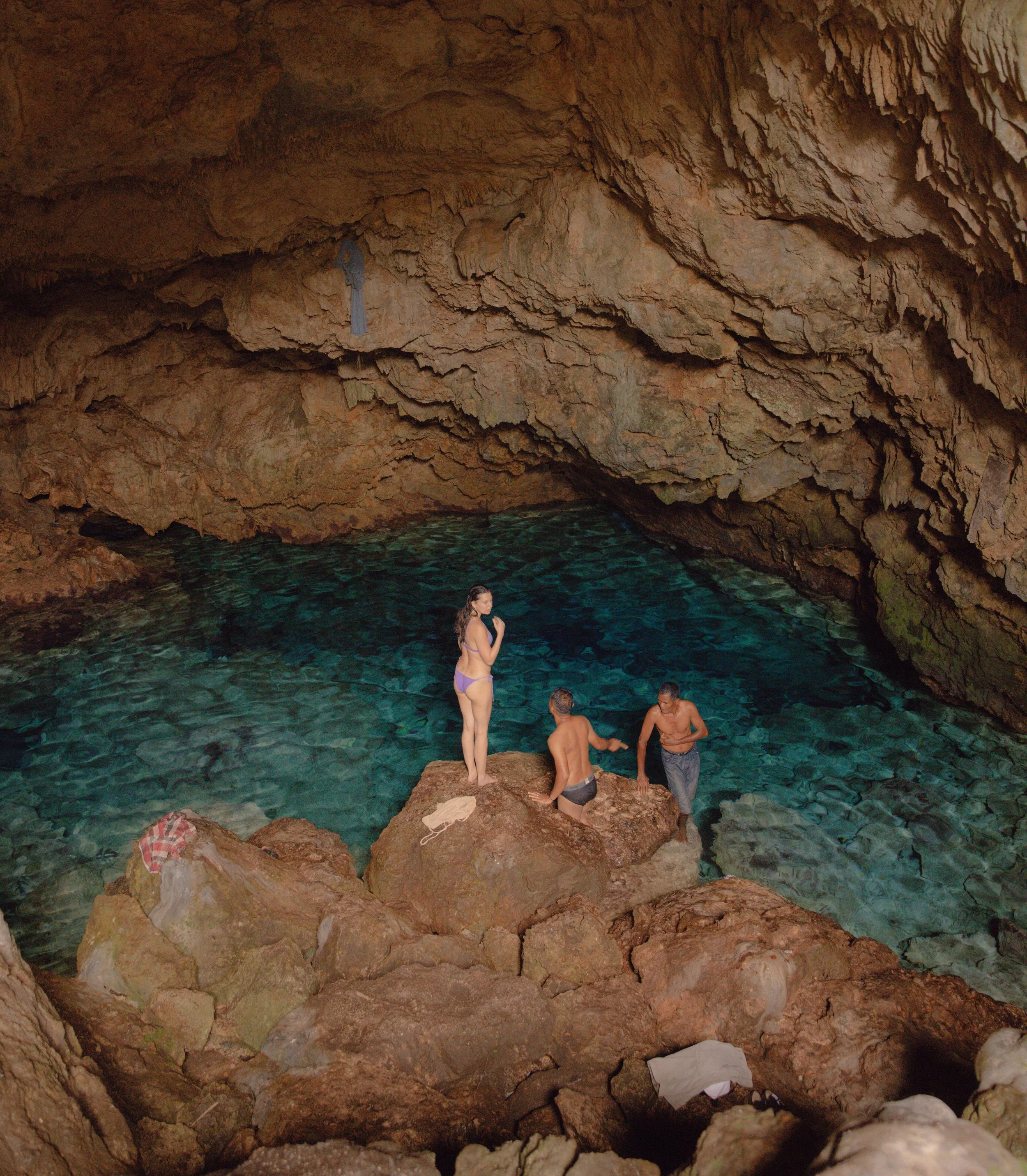People swimming and relaxing in a natural underground cave pool with rocky walls and a high ceiling.