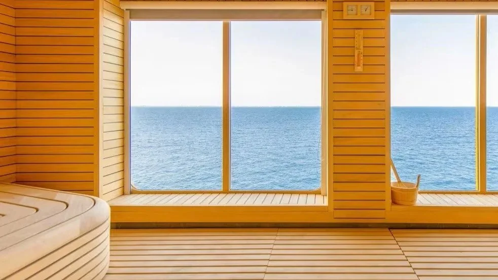 View of the ocean through large windows in a wooden room, with a sauna bench on the left and a bucket with a ladle on the right.