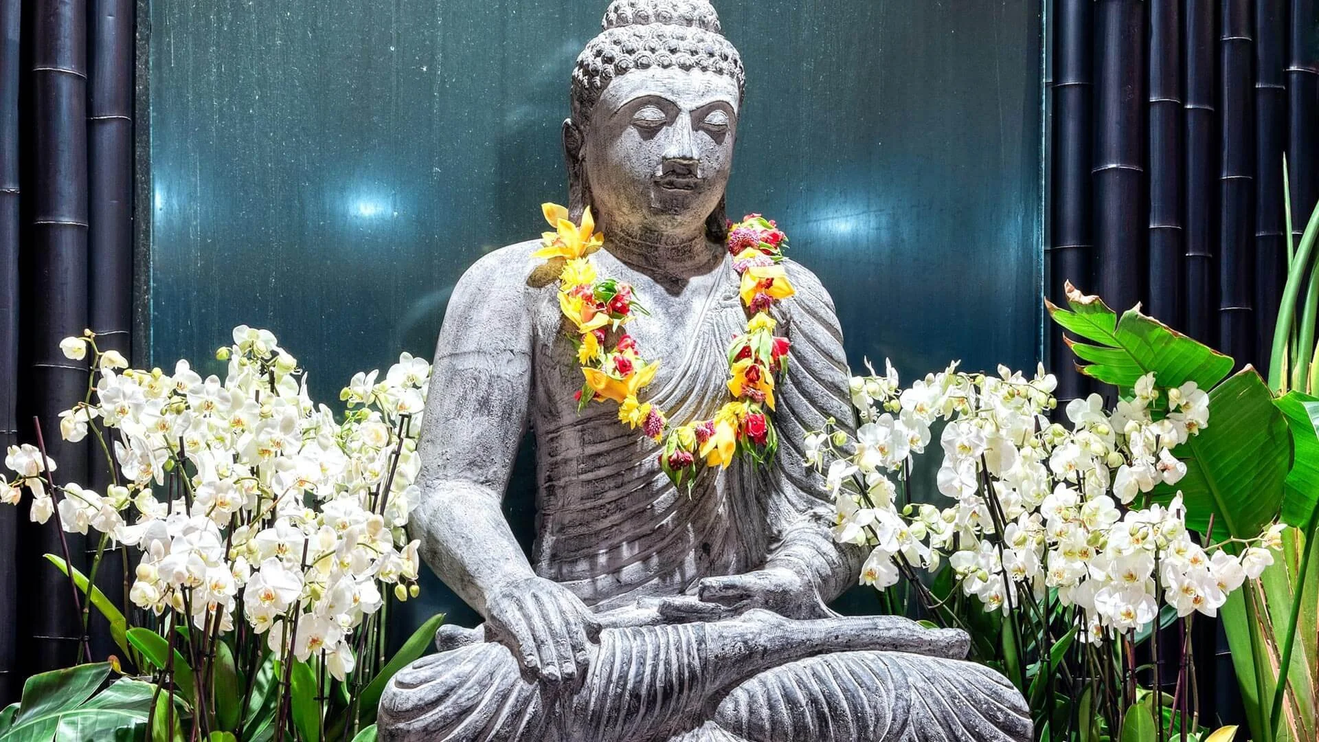 Statue of a meditative figure adorned with a colorful flower lei, surrounded by white orchids and green foliage.