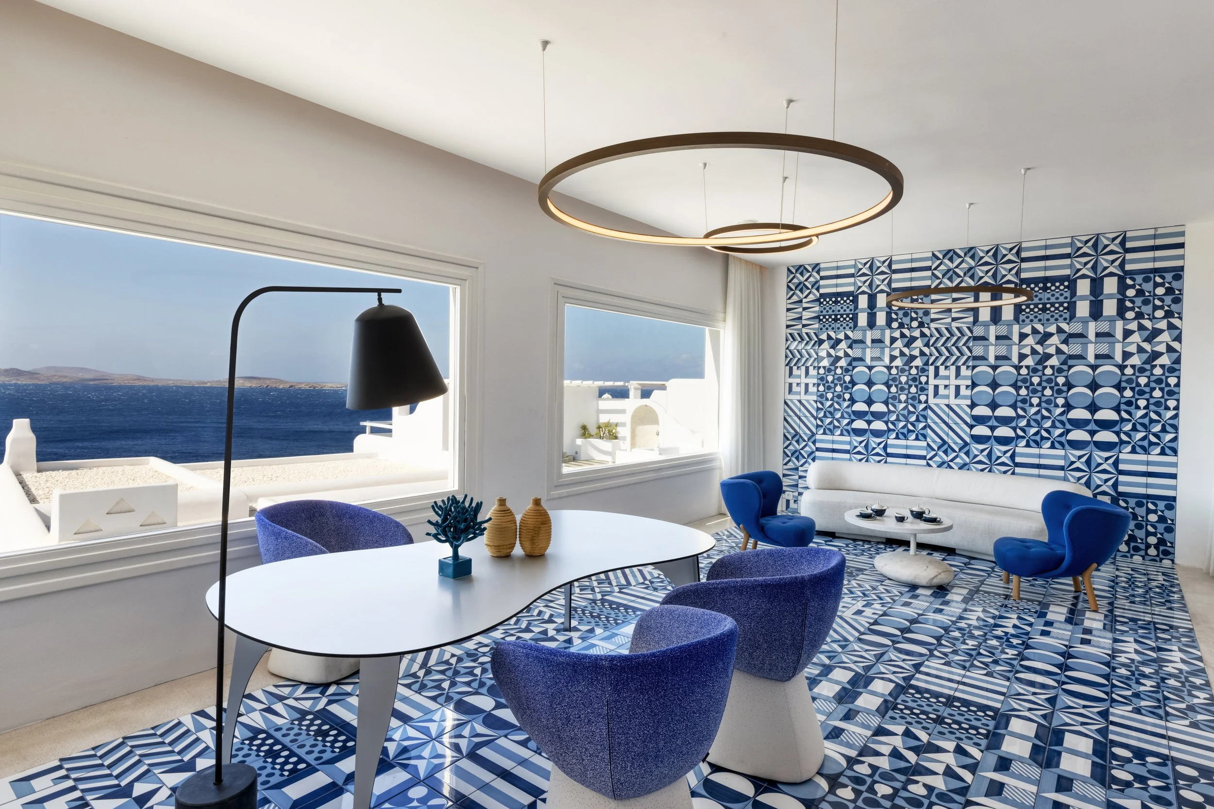 A room with blue and white decor, large windows showing a view of the sea, patterned blue and white tiles, a white curved sofa, blue armchairs, a white oval dining table with blue chairs, and unique pendant lighting fixtures.