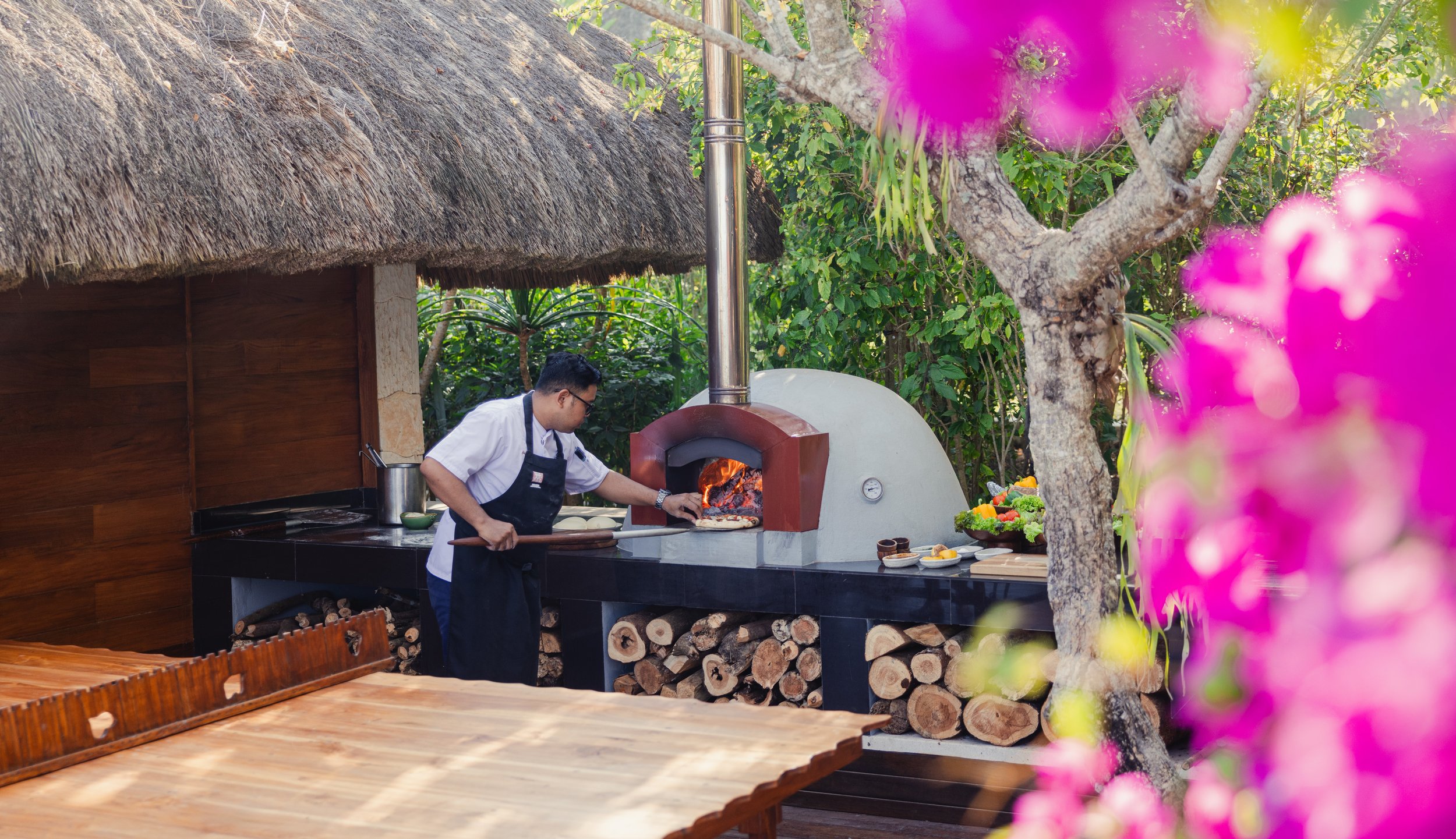 A chef using a pizza peel to remove a pizza from a wood-fired outdoor pizza oven in a garden setting, with a thatched roof structure and pink flowers in the foreground.
