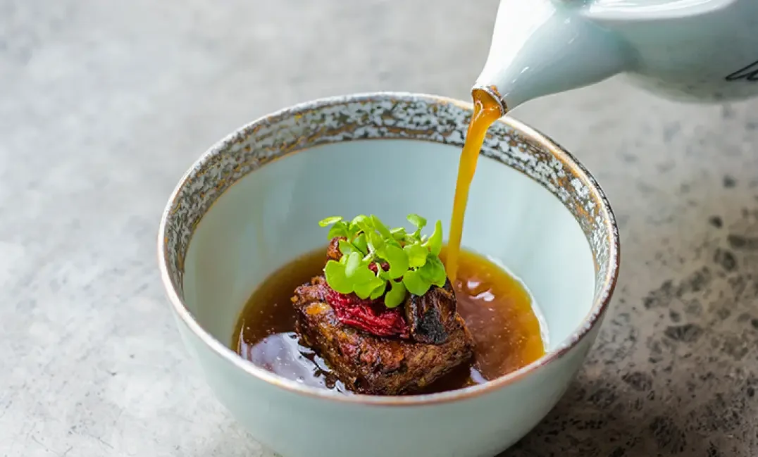Pouring gravy over a piece of meat topped with greens in a bowl.