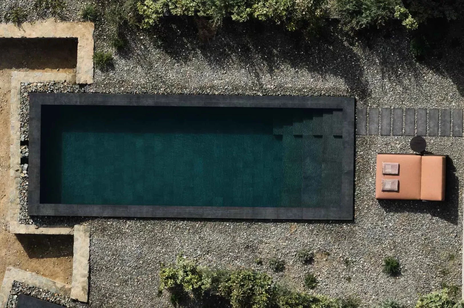 A rectangular outdoor swimming pool with a dark border, surrounded by gravel and a tiled area with a bench and a small black round object, viewed from above.