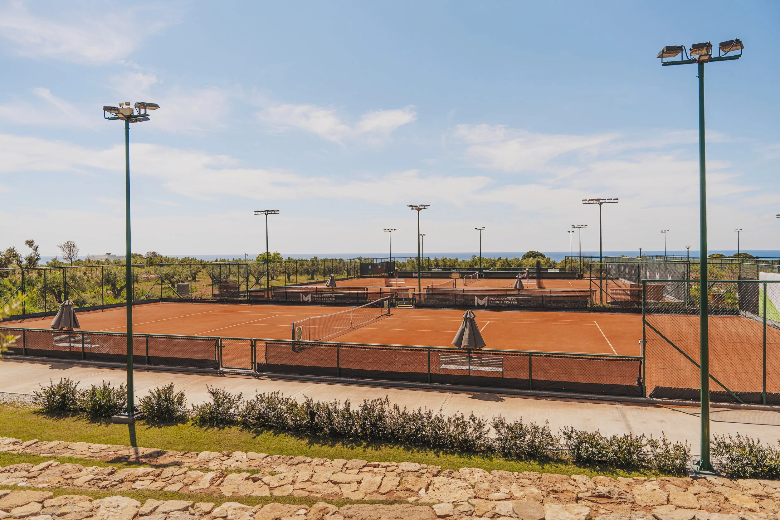 Multiple empty outdoor clay tennis courts surrounded by a chain-link fence, with a blue sky and distant ocean in the background.