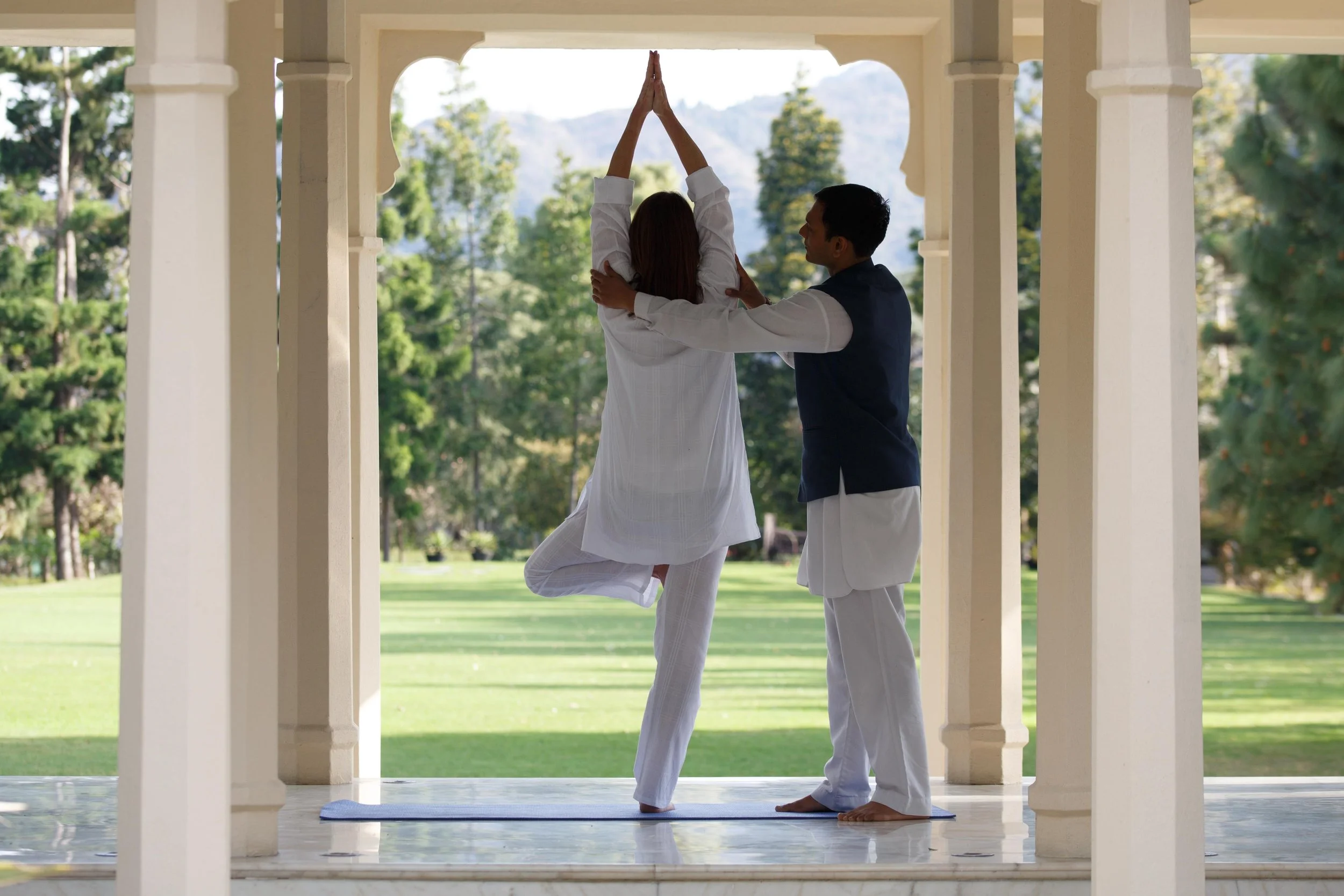 A woman and man practicing yoga together on a mat outdoors under a pavilion, surrounded by green grass and trees.