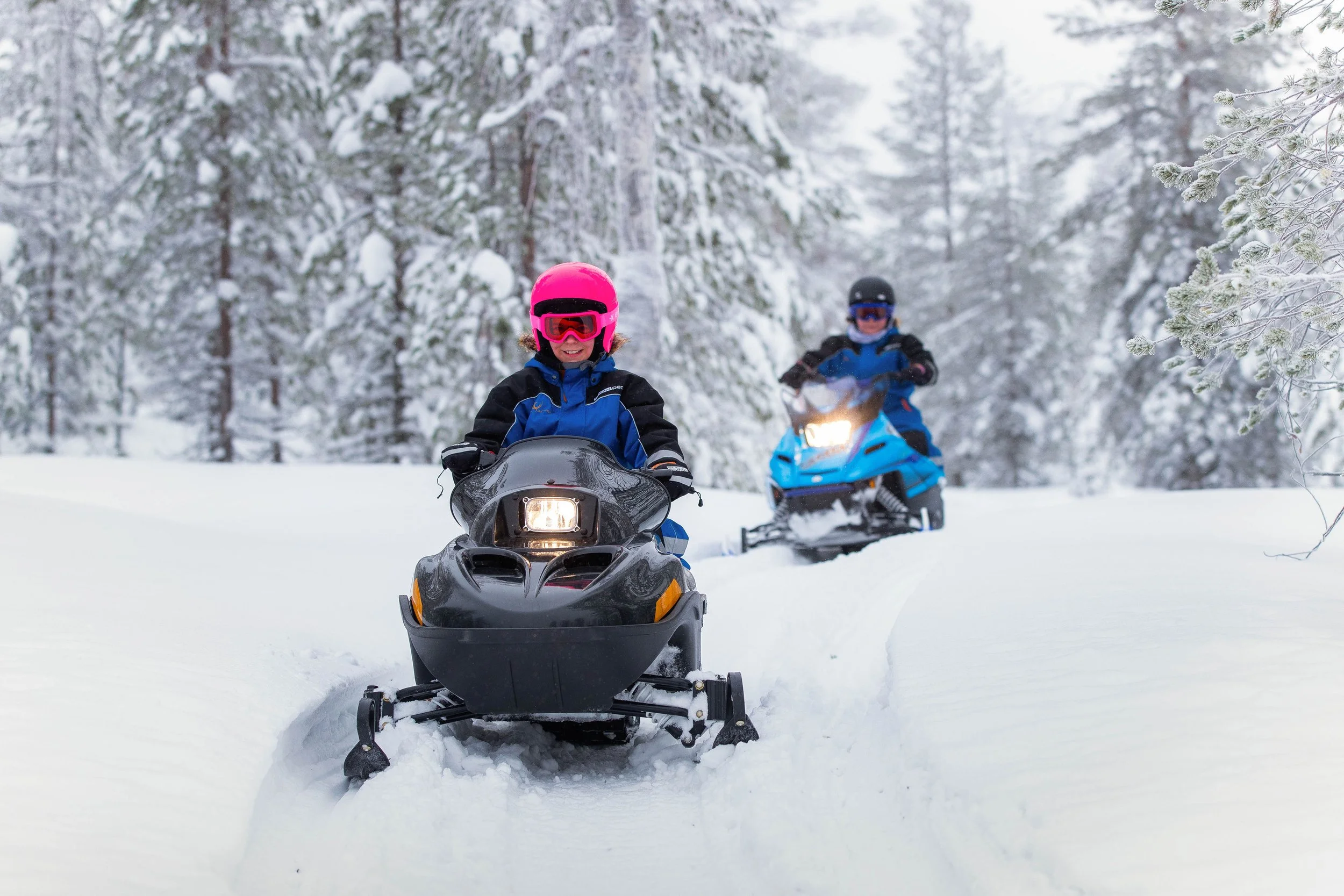 Two children snowmobiling in a snowy forest, wearing helmets and winter gear.