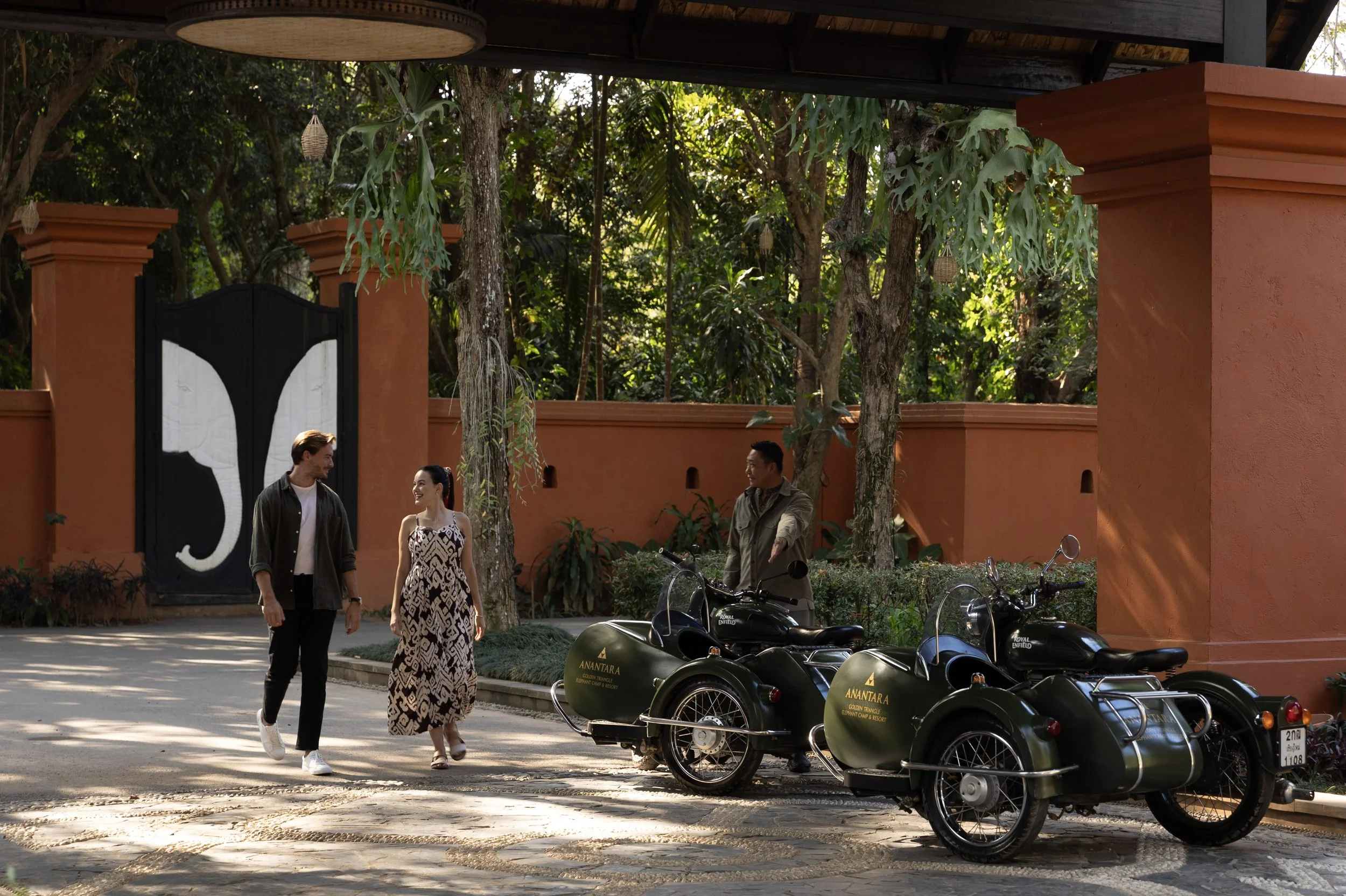 Two couples walking and talking near vintage sidecar motorcycles in front of an orange wall with a large black and white gate and lush trees.