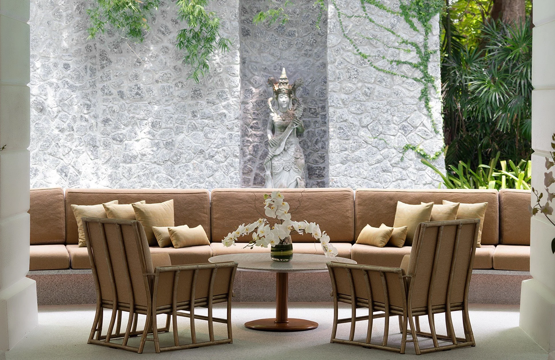 Indoor seating area with beige sofa, six beige pillows, four rattan chairs around a small round table with a vase of white orchids, and a stone wall with a statue and greenery in the background.