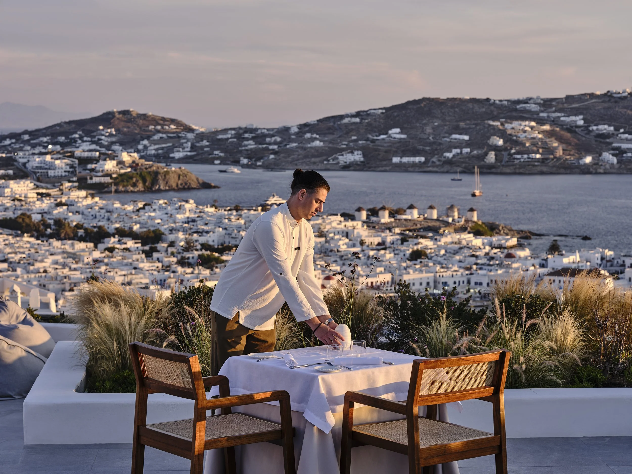 A waiter setting a table outdoors with a scenic view of a coastal town at sunset, with white buildings, water, and hills in the background.