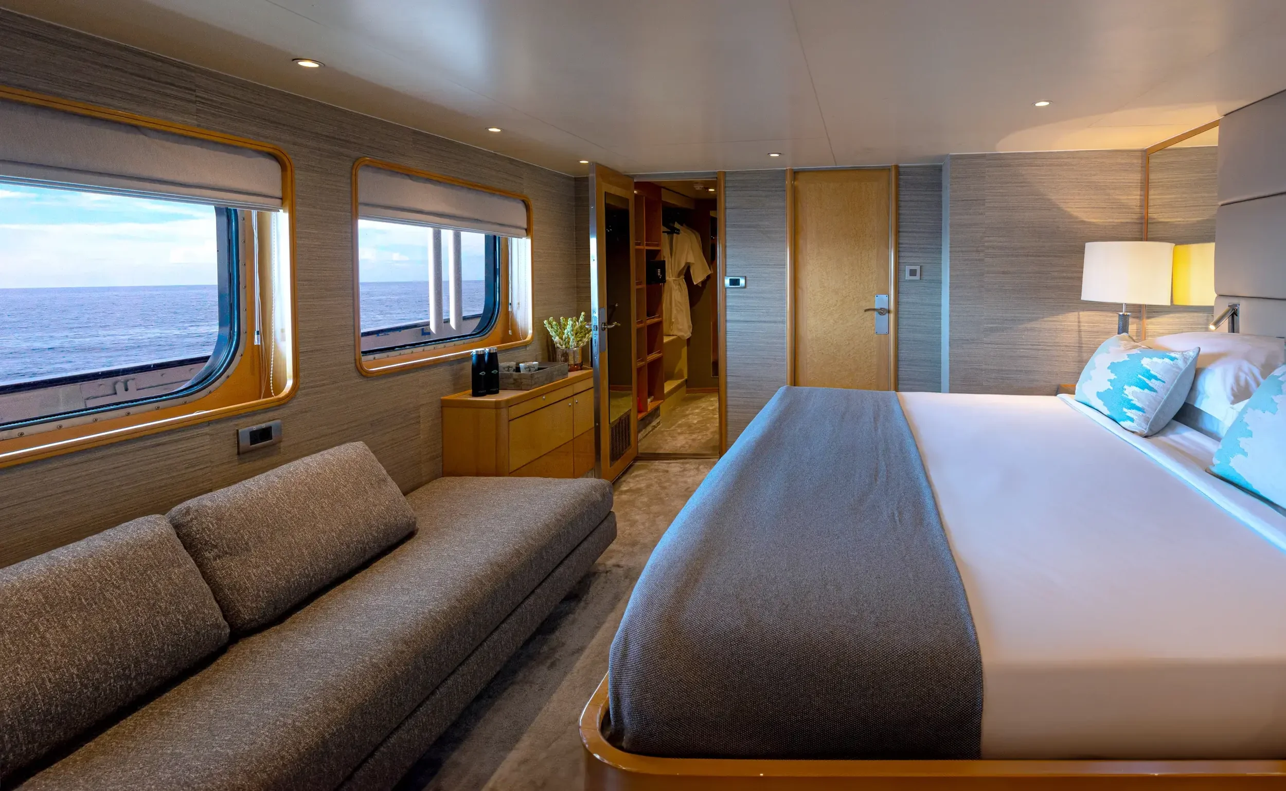 Interior of a cruise ship cabin with a large bed, a sofa, two windows showing the ocean, and an open closet.