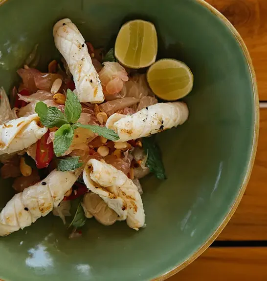 A green bowl with grilled squid, lime wedges, chopped onions, tomatoes, fresh herbs, and pine nuts.