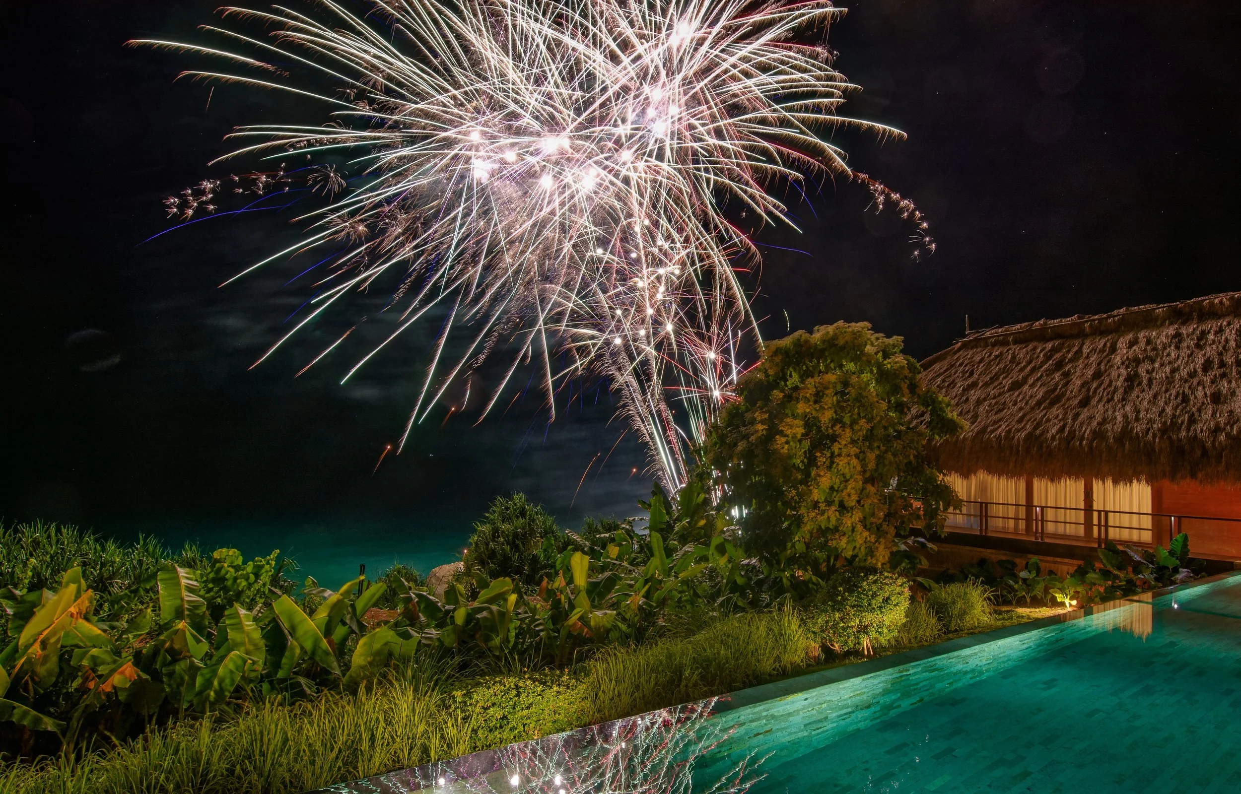 Fireworks display in the night sky over a tropical resort with a thatched roof building, lush greenery, and an infinity pool reflecting colorful fireworks.