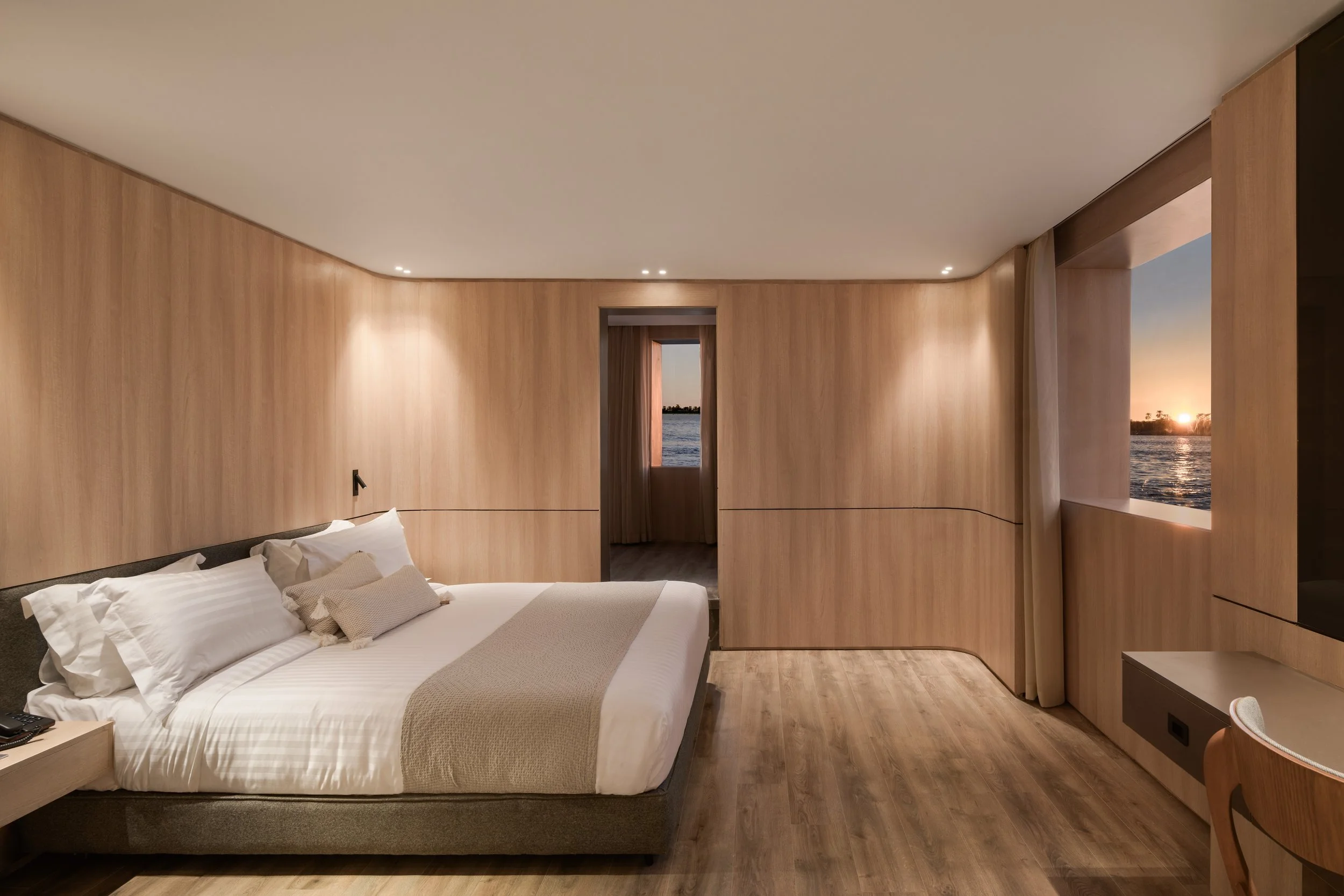 Modern room with a bed, wooden wall panels, and a view of a sunset over water through a window and door.