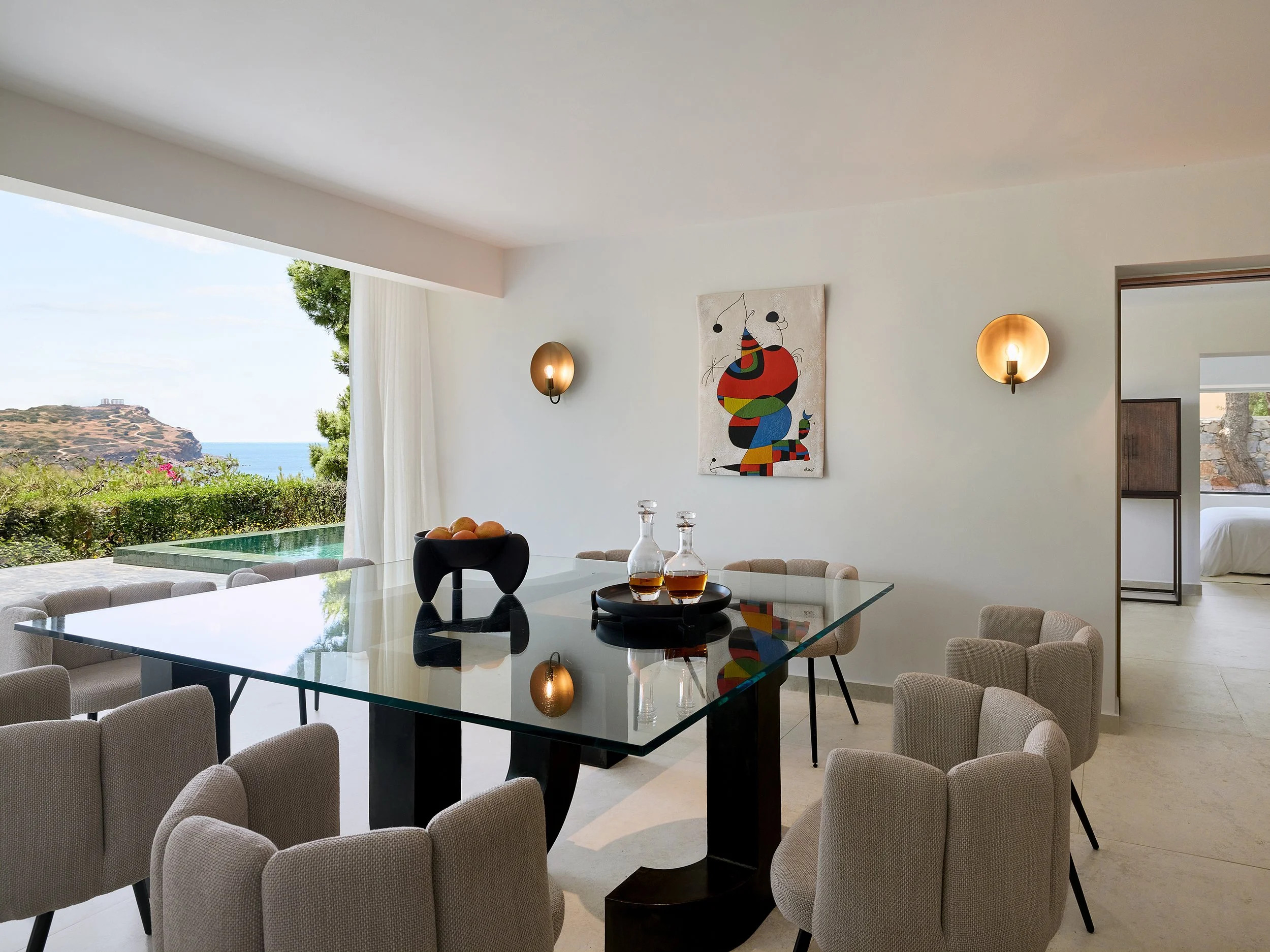 Modern dining room with glass table, beige chairs, colorful abstract art on white wall, outdoor view of vegetation, ocean, and a cliff, bedroom visible through doorway.