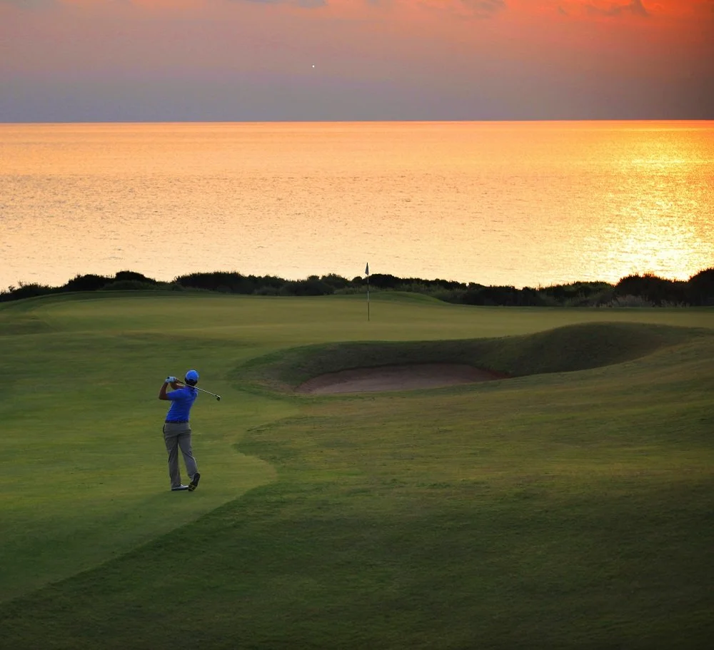 A golfer taking a swing on a golf course at sunset with the ocean in the background.