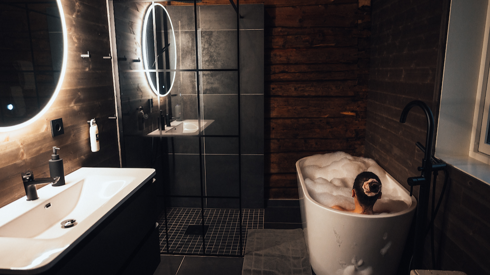 Person relaxing in a bubble bath in a modern bathroom with wood-paneled walls and illuminated round mirrors.