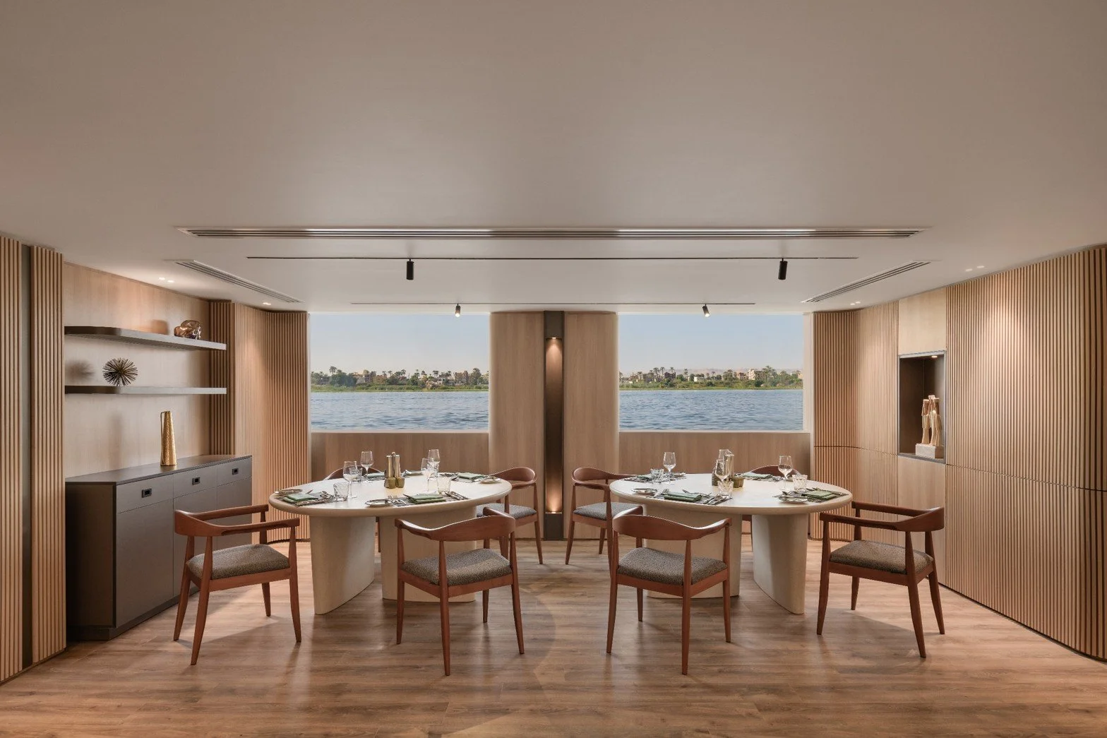Modern dining room overlooking the water with two round tables set with glassware, silverware, and napkins, and wooden chairs. The room features wood-paneled walls, a display shelf, and a view of the water and distant landscape through large windows.