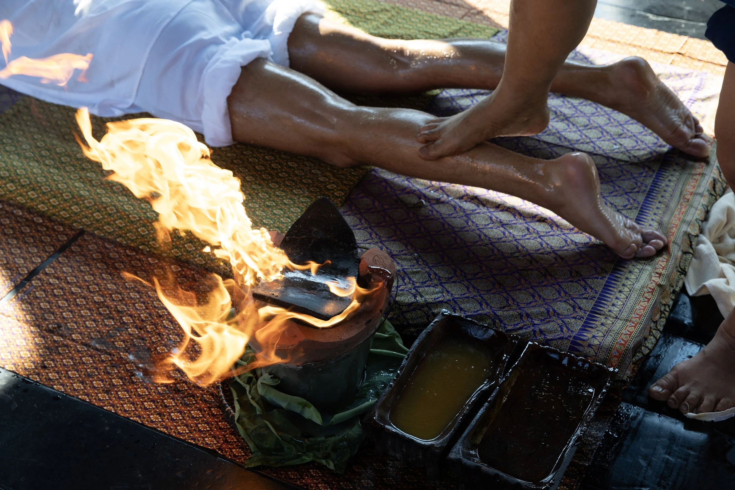A person receiving a traditional Thai massage while lying face down on a patterned mat, with an open flame near their legs as part of the ritual, and massage oils and tools nearby.