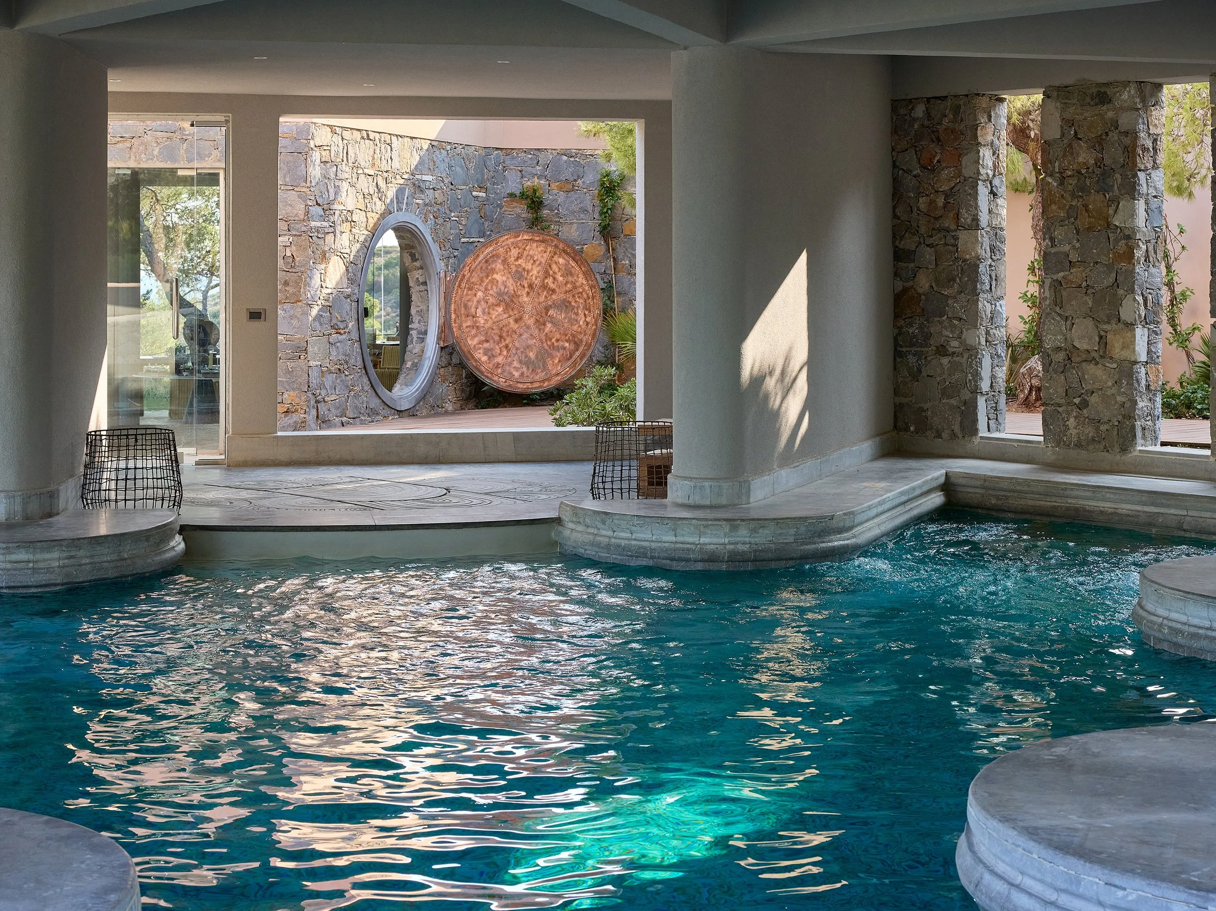 Indoor swimming pool with stone columns and walls, and an outdoor area with a stone wall featuring a large round mirror and circular decor.