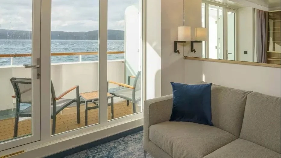 Living room with a beige sofa with a navy blue pillow, glass sliding doors leading to a small balcony, and a view of water and hills outside.