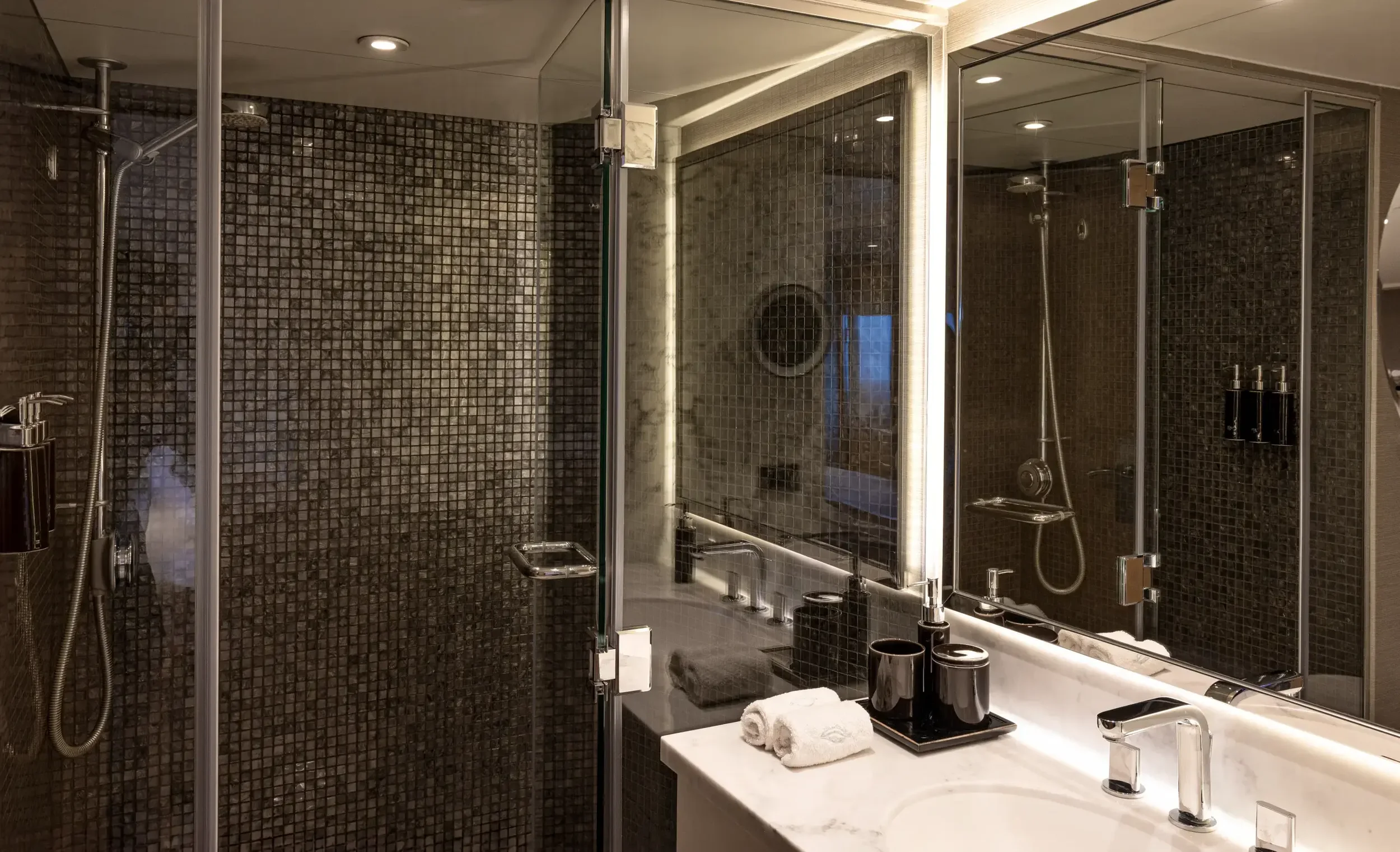 Modern bathroom with a glass-enclosed shower with dark mosaic tiles, a marble countertop, and a large mirror with integrated lighting. The countertop has rolled towels.