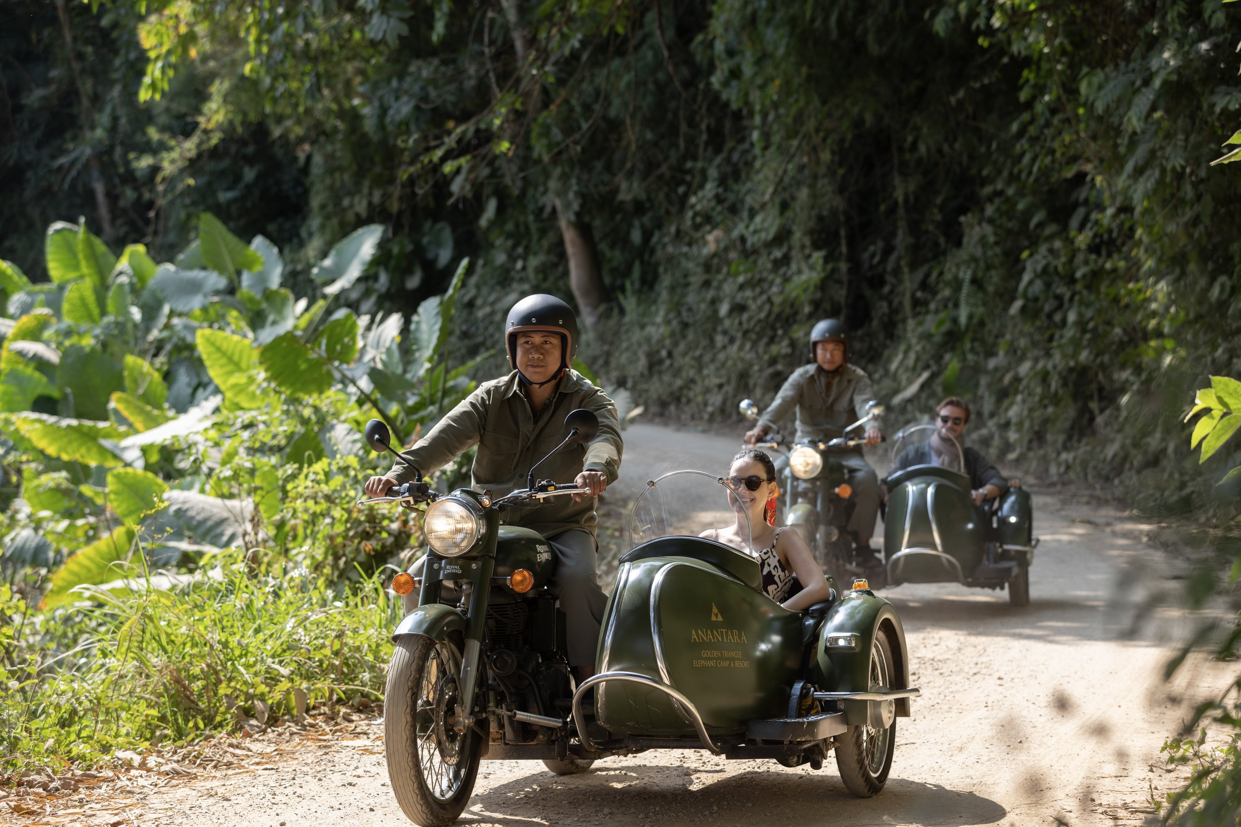 People riding motorcycles with sidecars on a dirt path through a lush green forest.