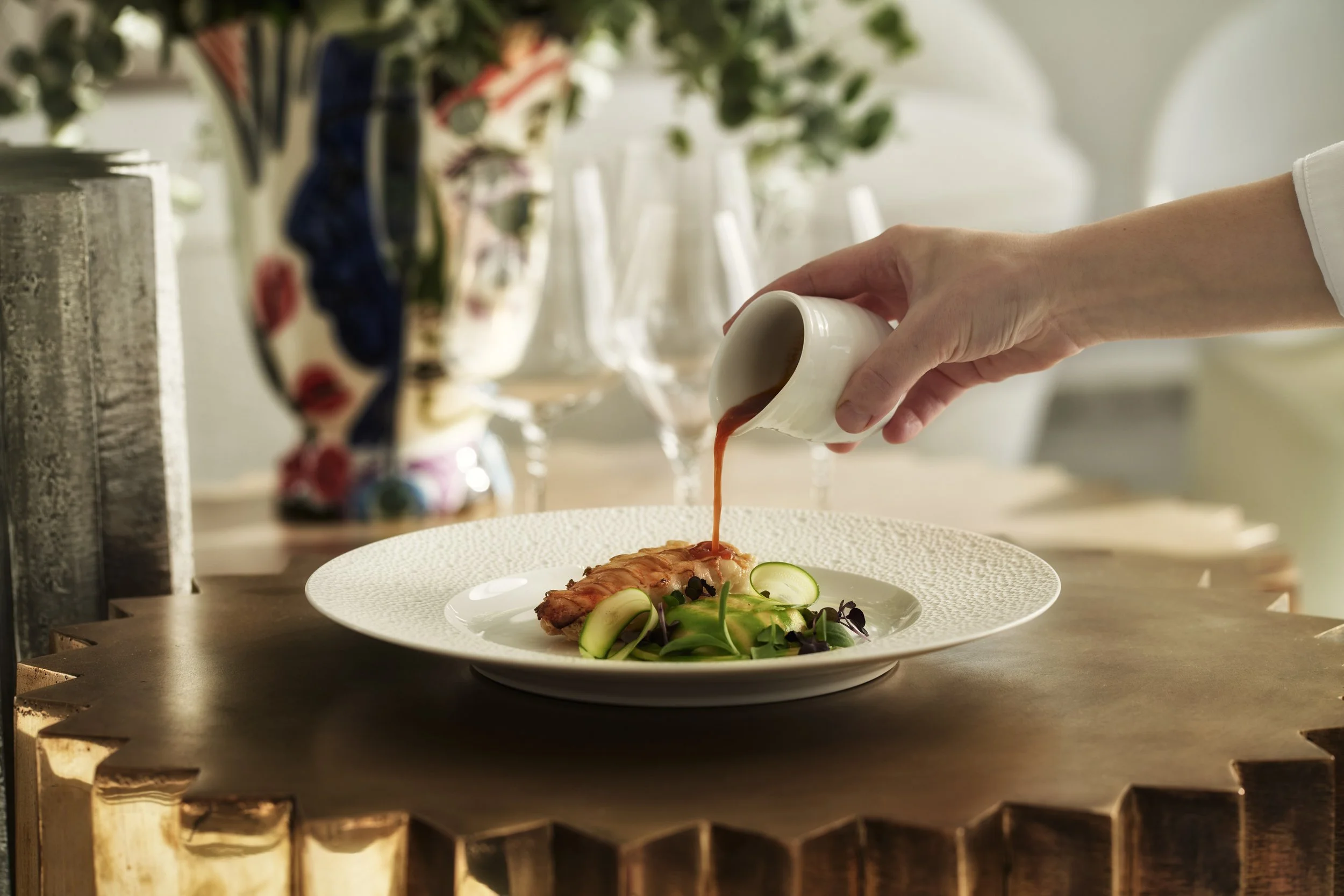 A person is pouring sauce over a plated dish of salmon and salad with cucumber slices on a table set for dining.