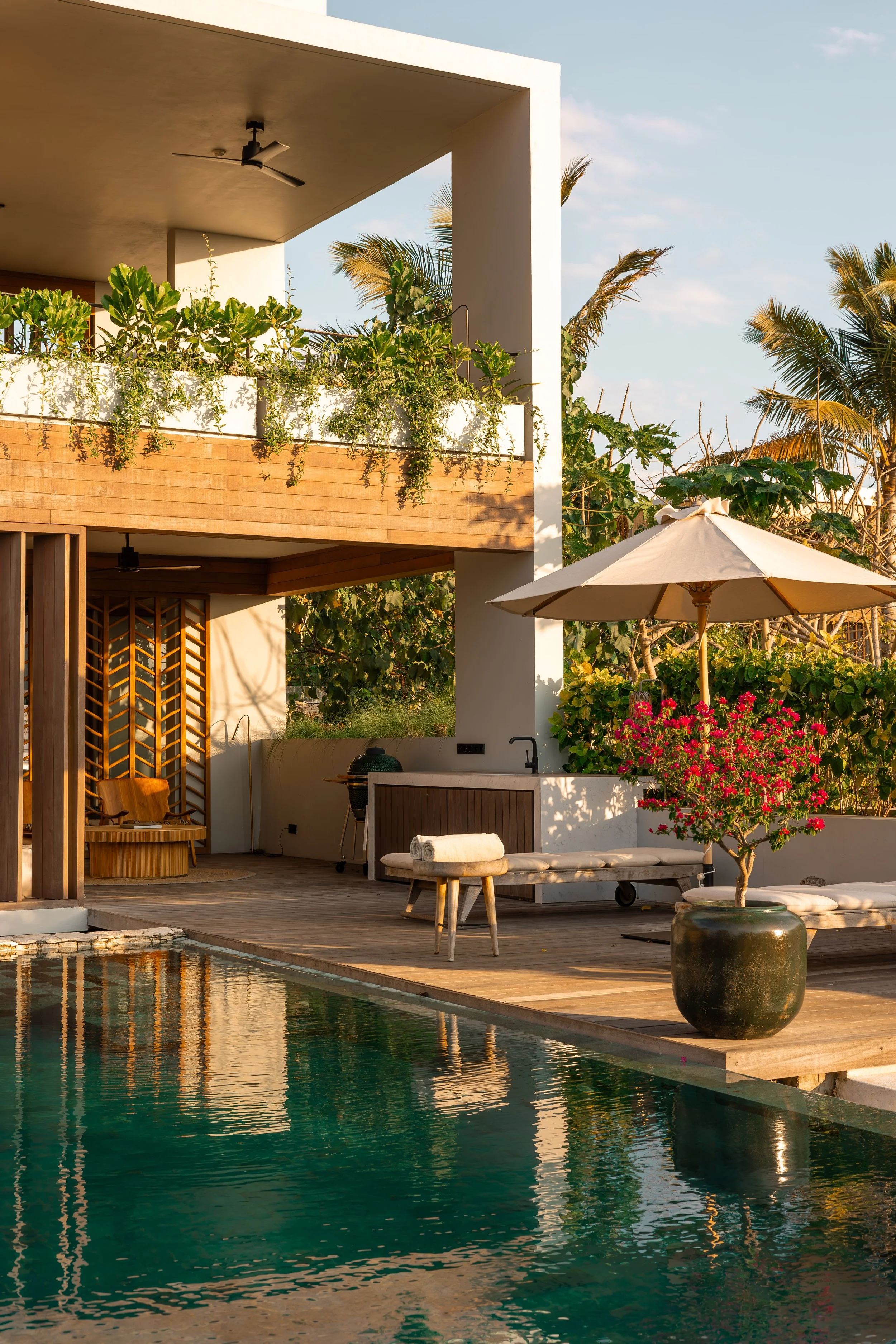A modern outdoor poolside area with a wooden deck, lounge chairs, large potted flowering plant, and an umbrella. In the background, there is a two-story building with white walls and greenery, including palm trees.