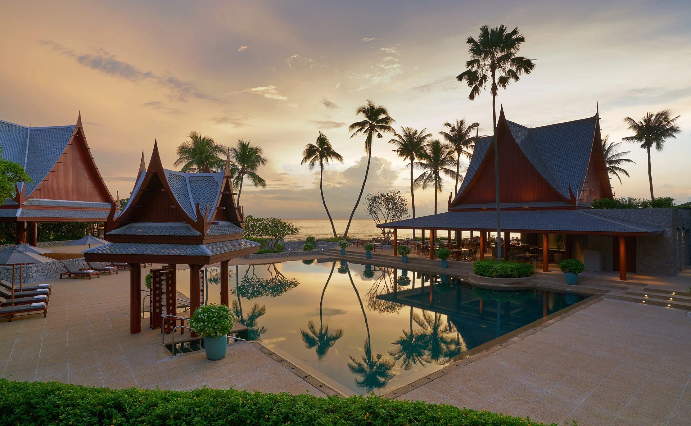 A tropical resort during sunset with traditional Thai-style buildings surrounding a swimming pool, palm trees, and a view of the ocean in the background.