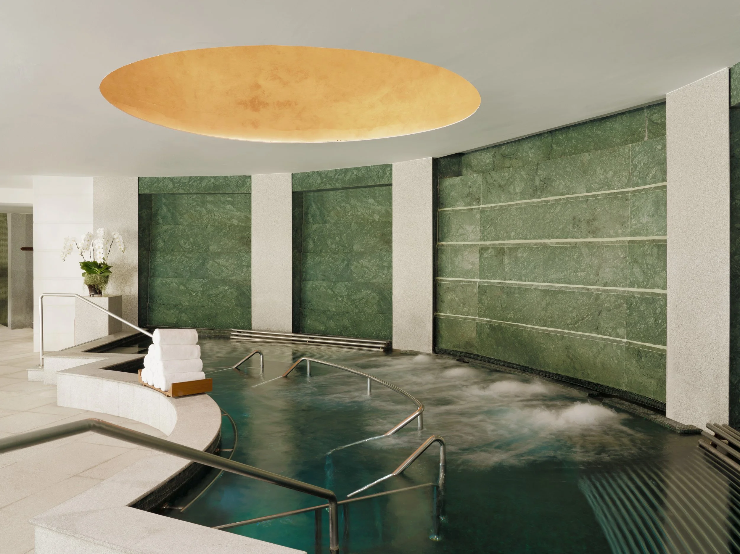 Indoor hot spring pool with stainless steel handrails, white rolled towels, a potted orchid, green marble walls, and a ceiling light fixture.