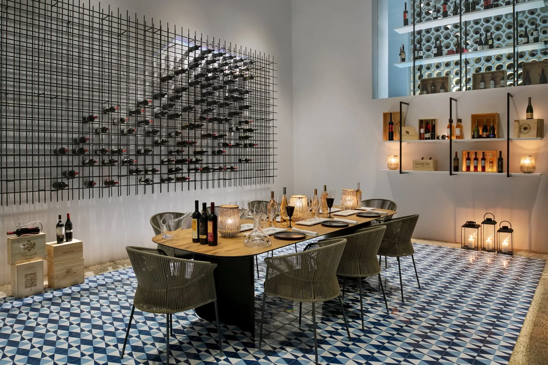 A wine-tasting room with a modern design. A long wooden table is set with wine bottles, glasses, candles, and tableware, surrounded by six chairs. On the left wall, there is a large black metal grid with numerous wine bottles stored horizontally. The right wall has shelves holding more wine bottles and decorative items, with candles in lanterns on the floor, creating a cozy ambiance. The floor features a geometric blue and white tile pattern.