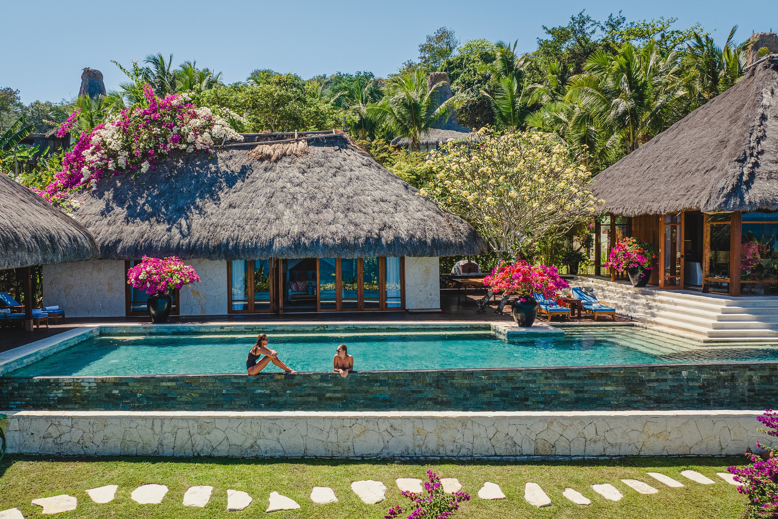 A tropical resort scene with a swimming pool, thatched roof huts, colorful flowers, lush green trees, and two women sitting in the pool enjoying the sunny weather.