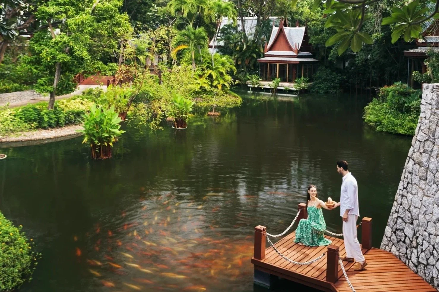A romantic scene by a lake with lush greenery and traditional Thai-style buildings in the background.