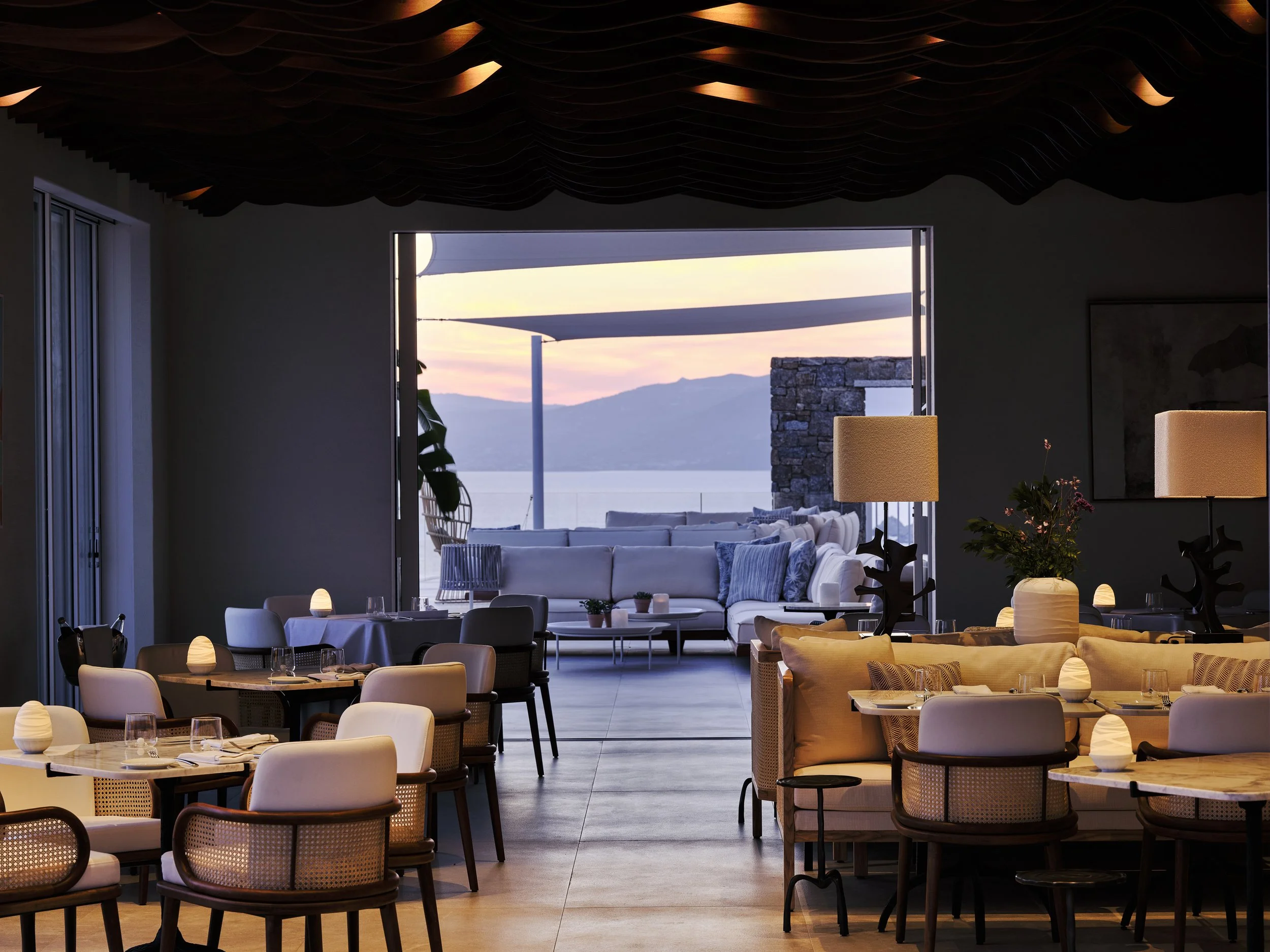 Interior of a restaurant with tables set for dining, featuring lamps and a view of a patio with outdoor seating, overlooking a lake and mountains at sunset.