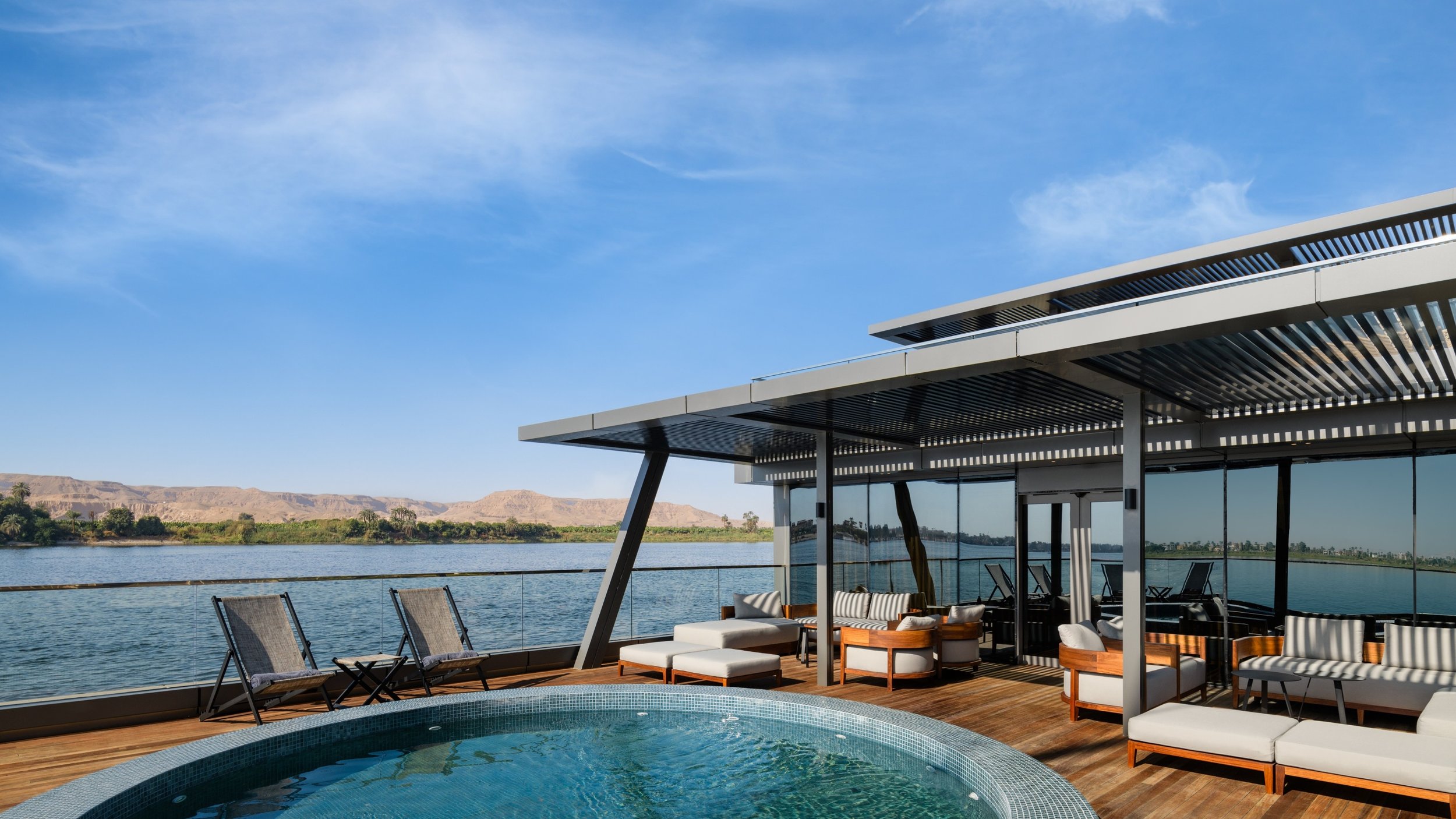 Luxury outdoor lounge with poolside seating overlooking the river and distant mountains under a clear blue sky.