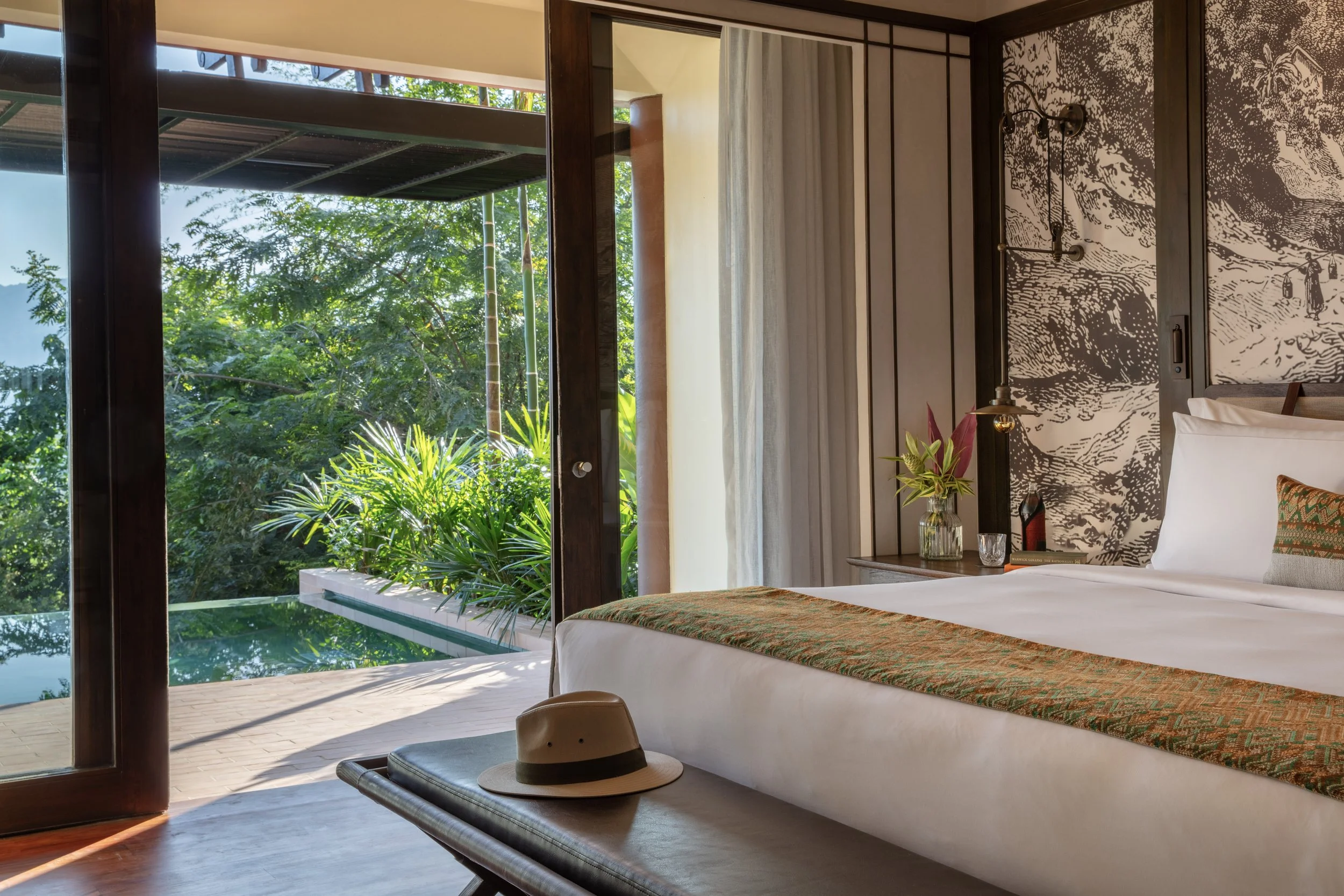 A bedroom with a large bed, decorated with a patterned runner, and a bench at the foot with a hat on it. The room has sliding glass doors opening to a balcony with lush green plants outside. There is a nightstand with a plant, a glass, and a lamp, and a decorative wall behind.