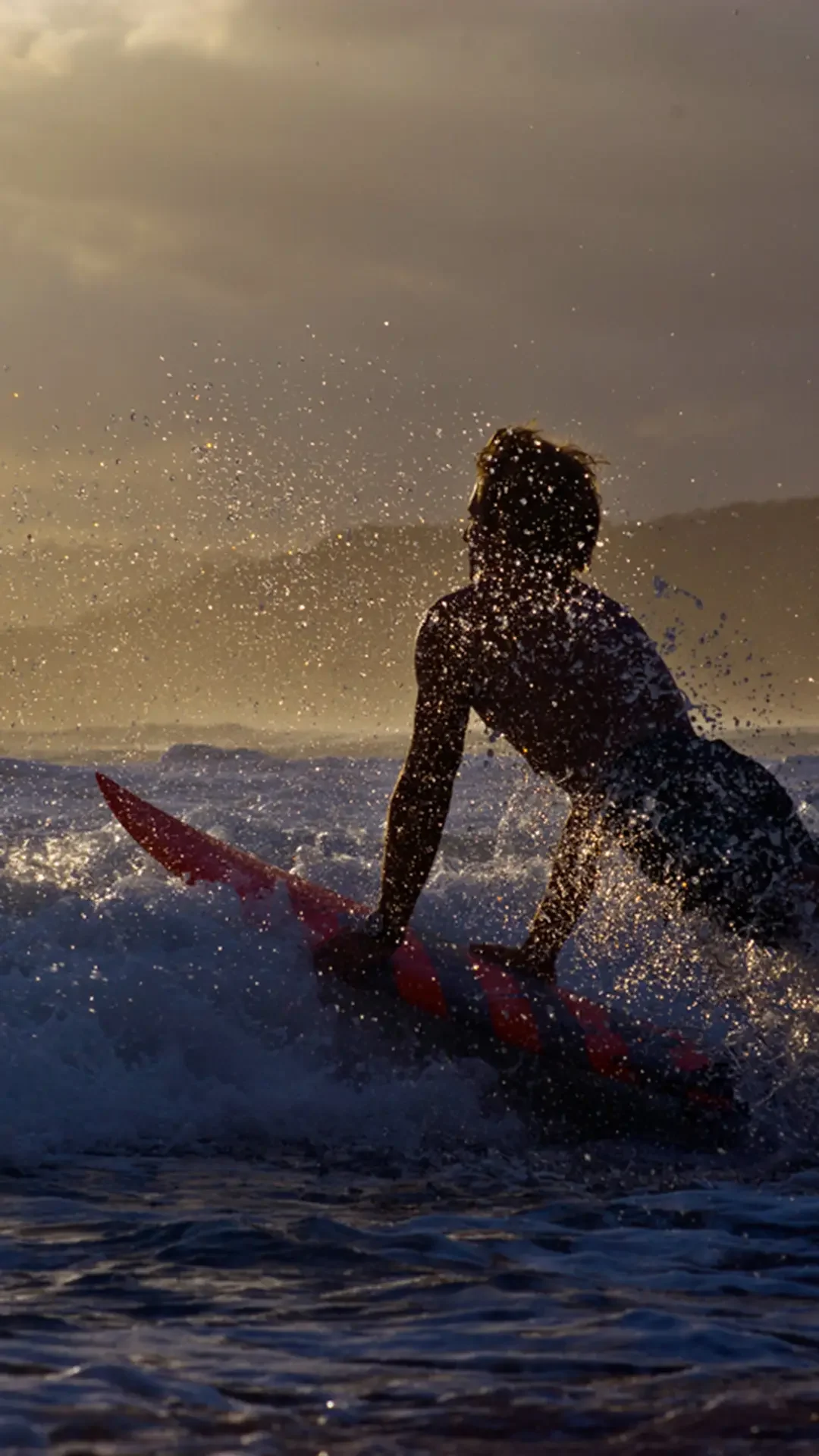 Silhouette of a person surfing on a red surfboard in the ocean during sunset or sunrise, with water splashing around and a cloudy sky in the background.