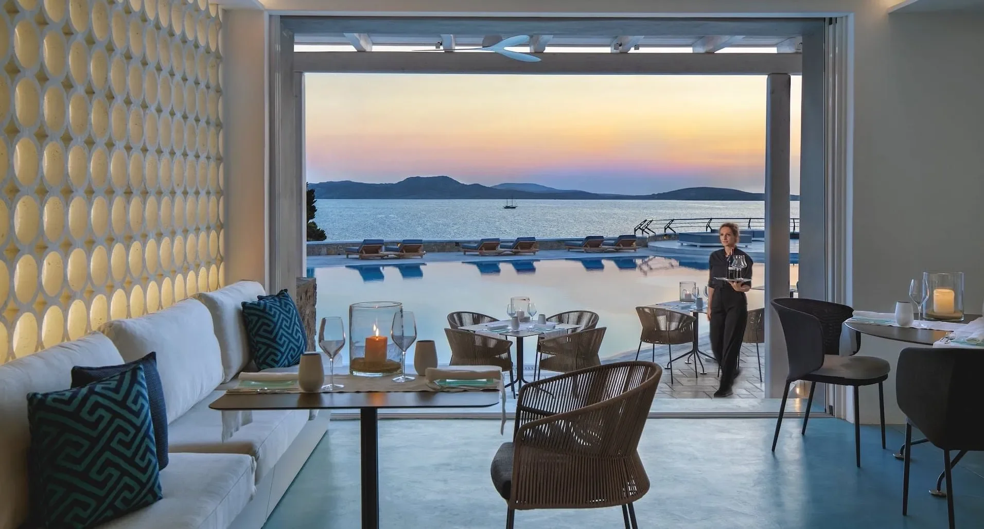 A cozy dining area with a view of an outdoor pool, ocean, and mountains at sunset. There are tables set with glasses, candles, and tableware, and a waiter is serving.