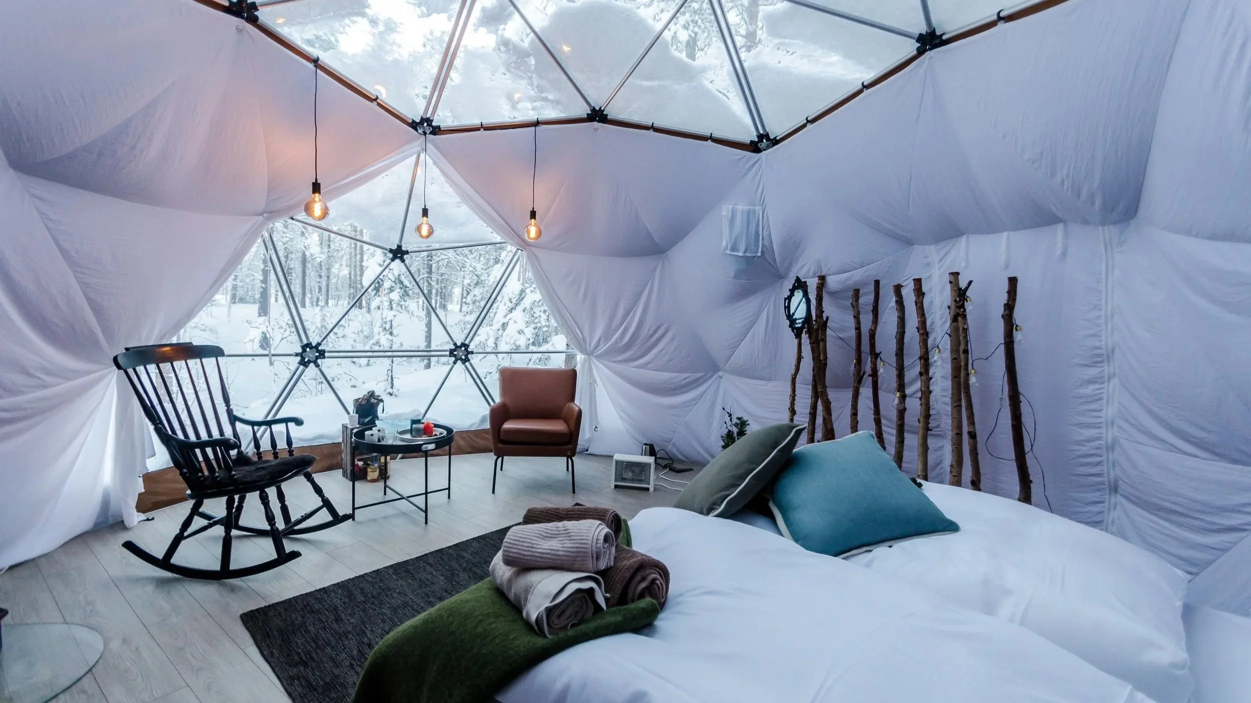 Cozy glamping tent interior with snow view, featuring a bed with rolled towels, pillows, a rocking chair, a small table, an armchair, hanging light bulbs, and decorative wooden sticks with fairy lights.