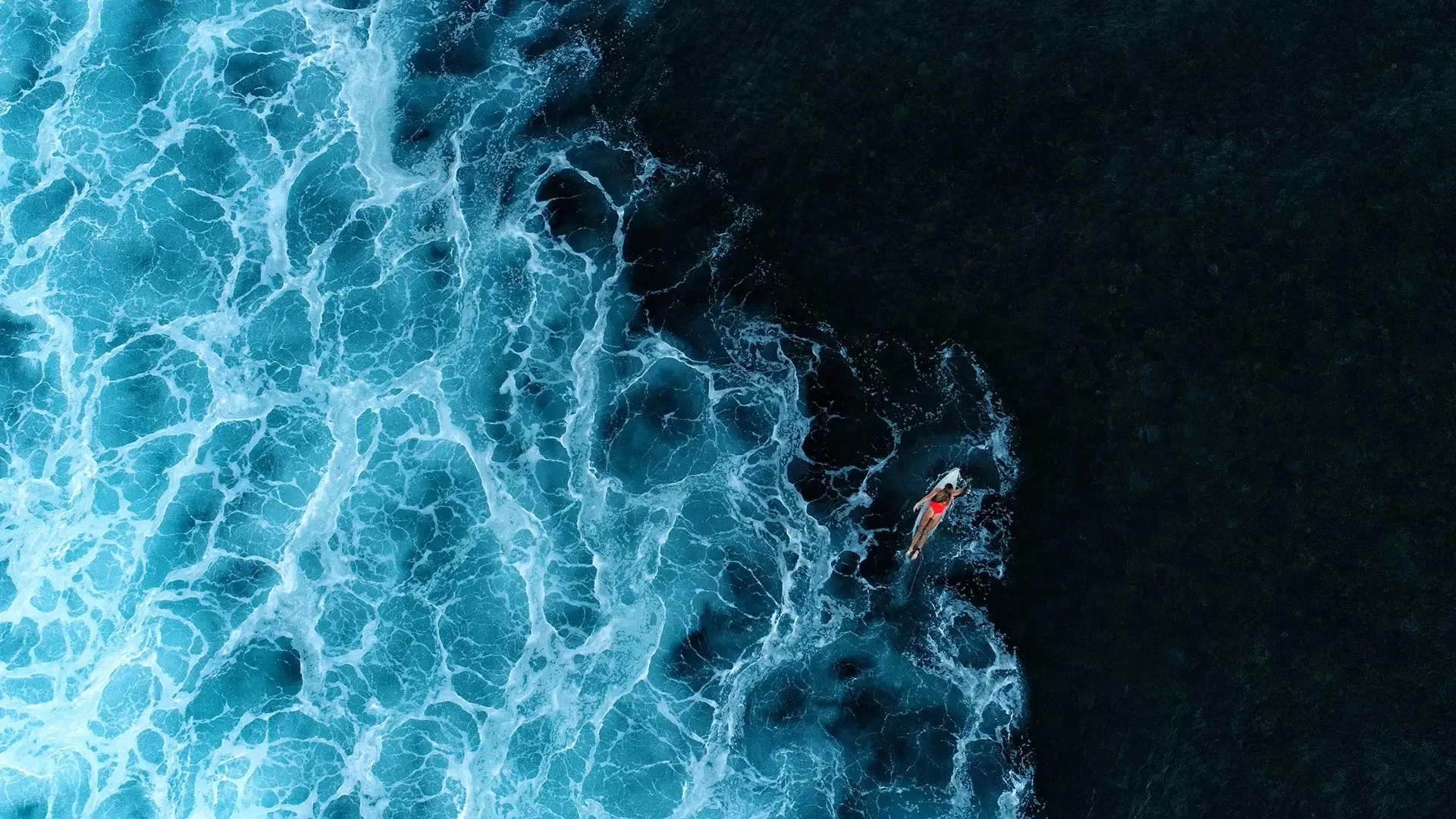 Aerial view of a person on a surfboard in the ocean, with white foamy waves on the left and dark water on the right.
