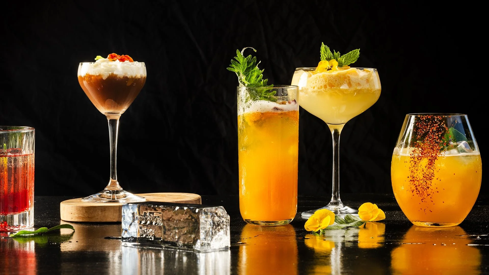 A variety of colorful cocktails on a dark surface, decorated with fruit, herbs, and ice cubes, with a black background.