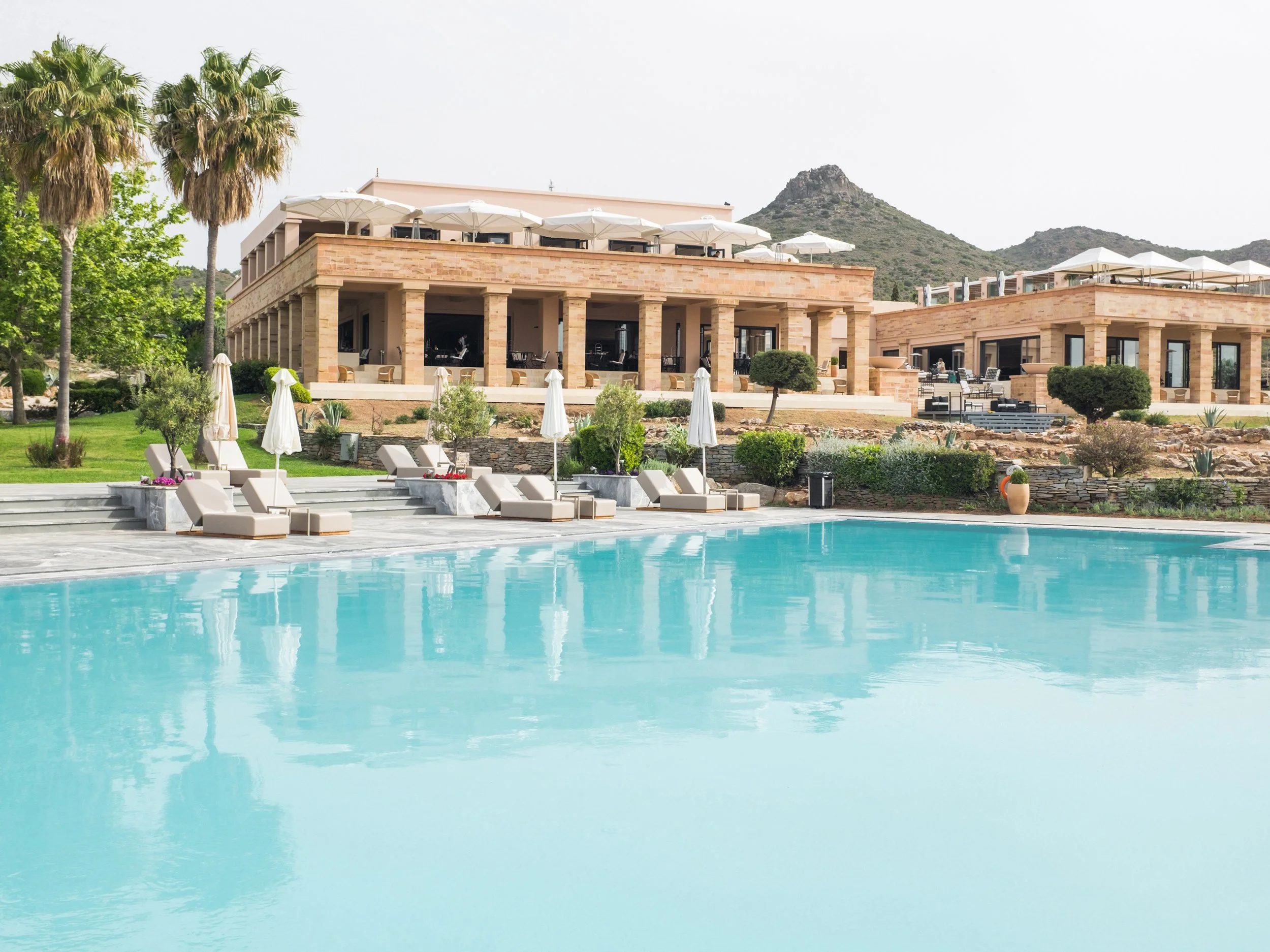 Luxury hotel with a large swimming pool, surrounded by lounge chairs and umbrellas, lush greenery, and a modern building with outdoor seating and a mountainous landscape in the background.